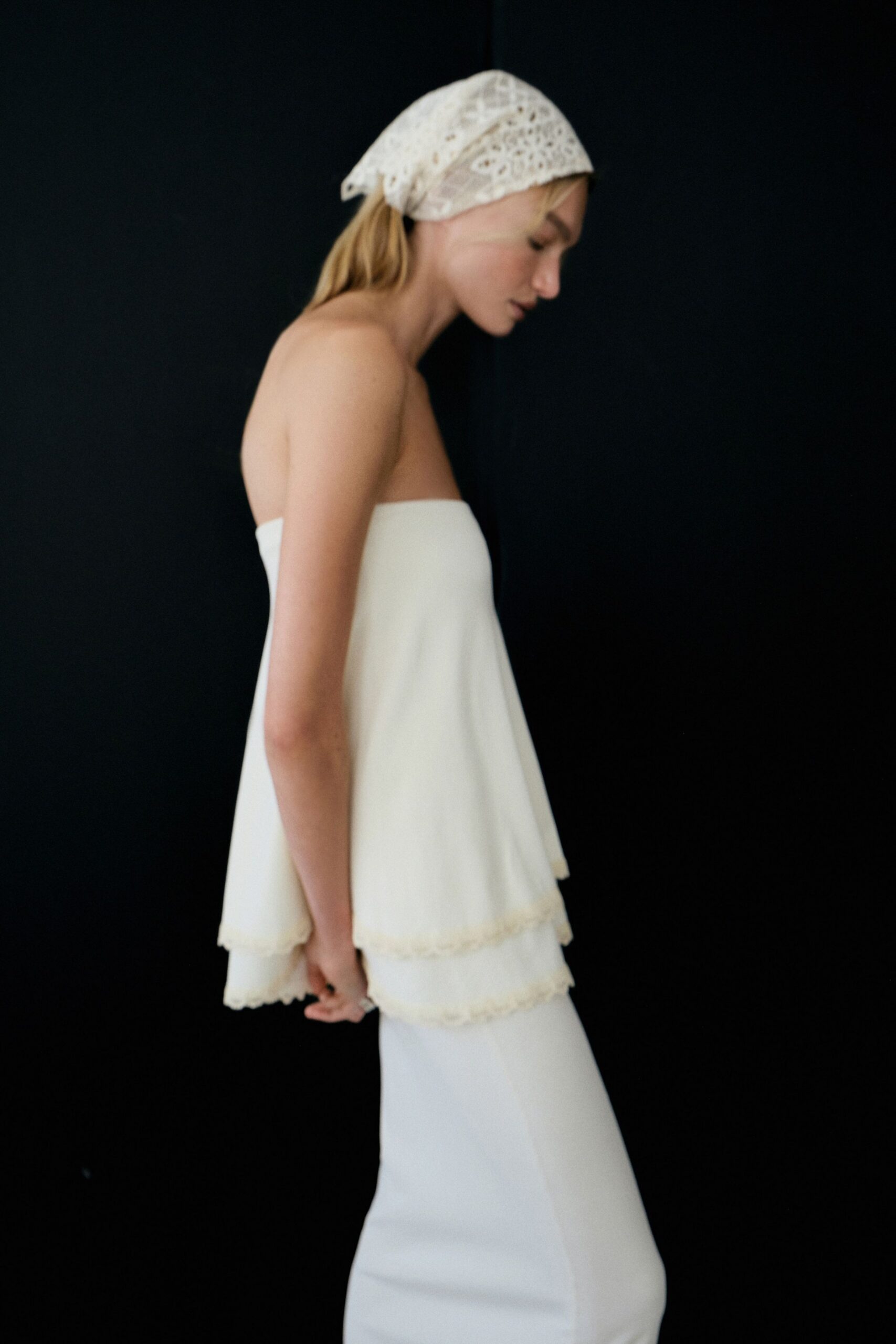 White eyelet kerchief tied low over a loose blond ponytail. Strapless ivory trapeze top in two floating tiers with tiny scalloped edges. Long ivory column skirt, clean and close through the hip. Sunday morning hotel curtains, still barefoot after washing out the salt.
