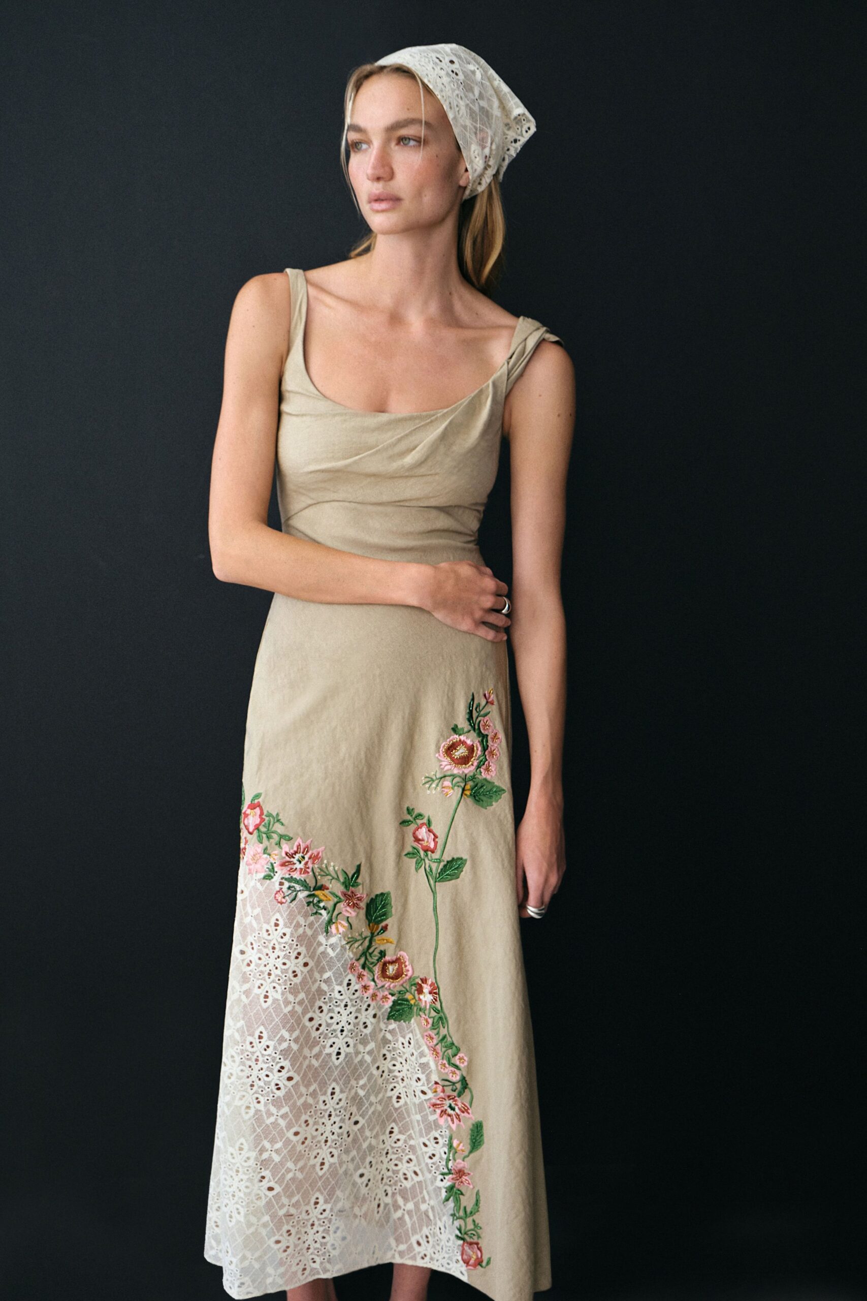 White eyelet kerchief tied close over the crown. Oat slip dress with twisted straps, floral embroidery climbing over cutwork lace hem. Stacked silver bands on both hands. Picking apricots behind the house before the sun reaches the fence.