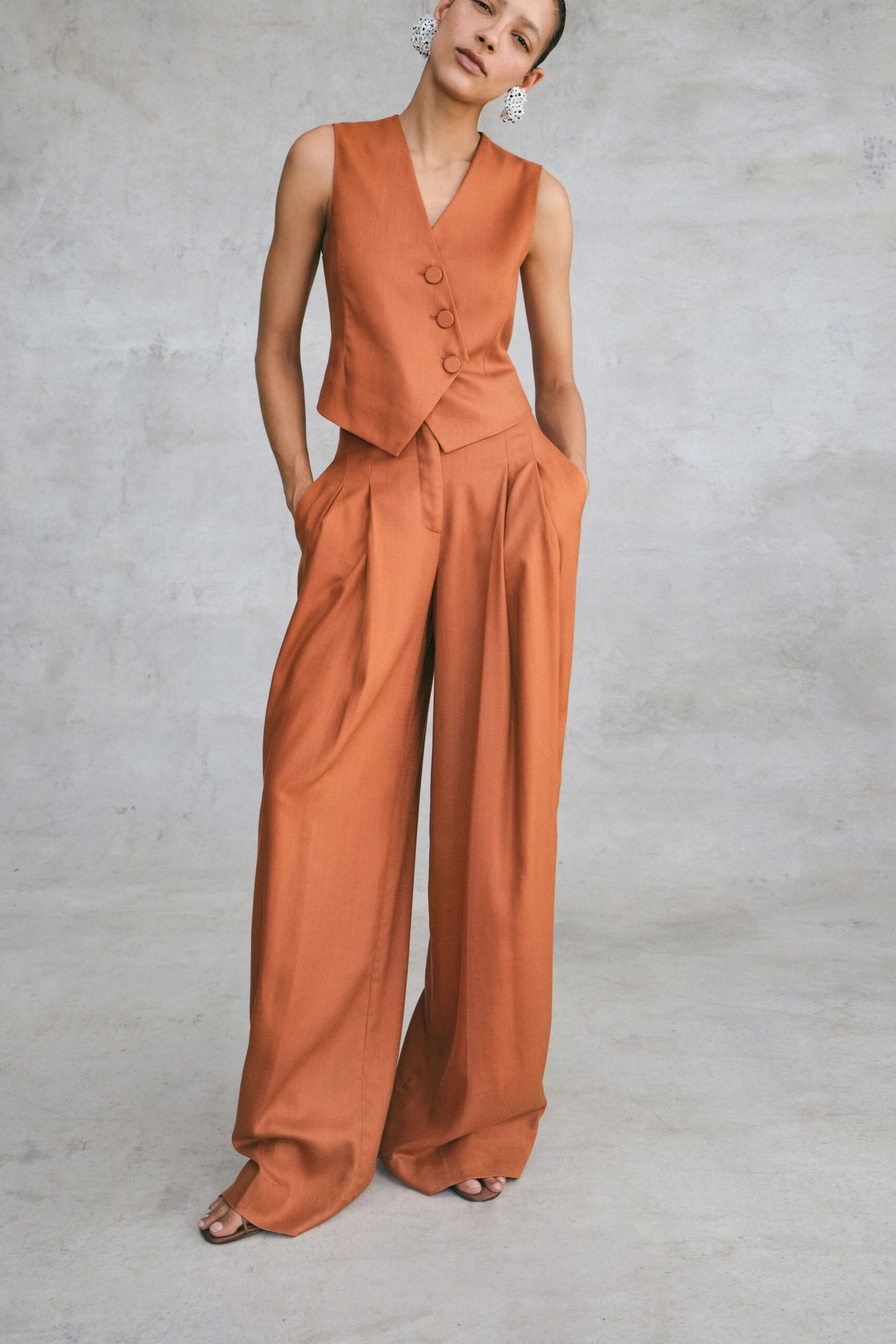 Clustered white bead earrings hanging almost to the jaw. Terracotta suiting vest with a deep V and three covered buttons. Matching wide-leg trousers, double pleats opening from the pockets. Flat brown thong sandals peeking from the pooled hem. For rooftop drinks before the heat lifts off the concrete.