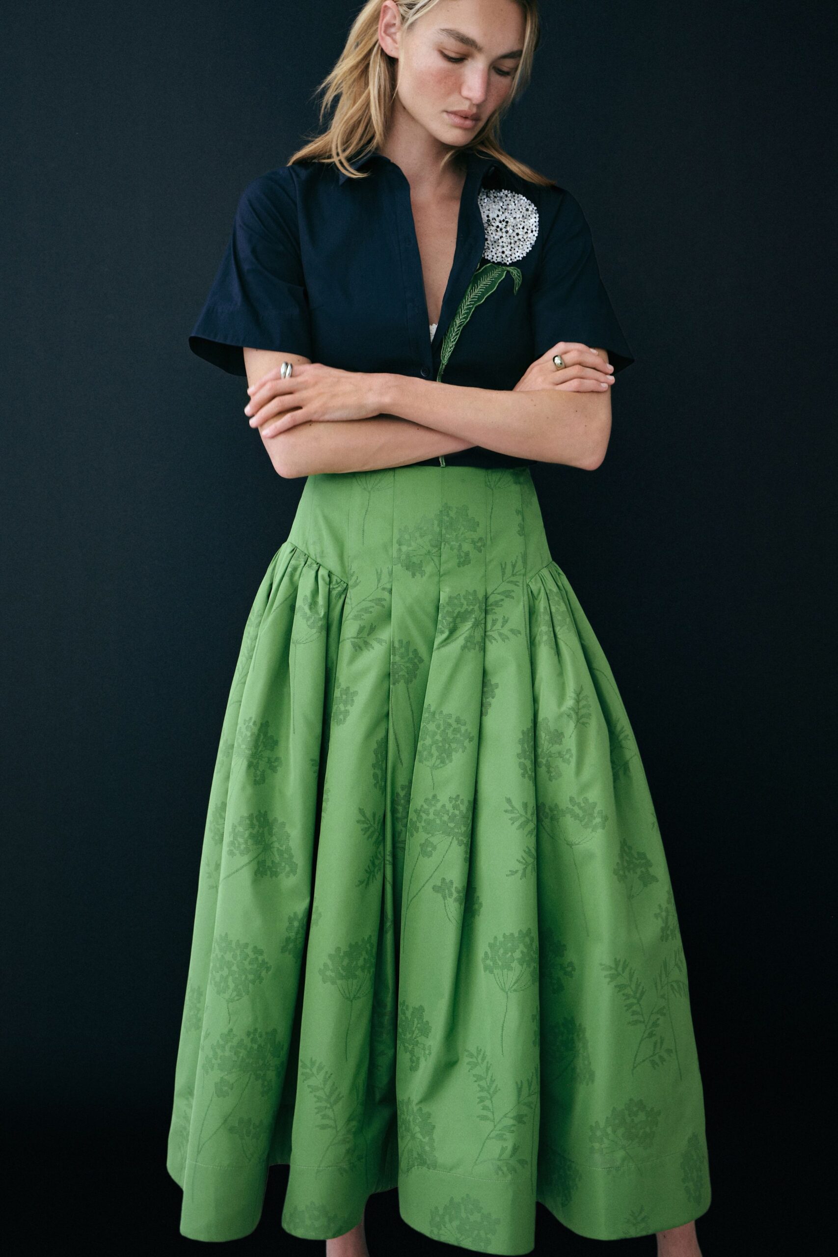 Navy short-sleeve shirt with a white beaded flower climbing one lapel. White triangle bralette peeking through the deep placket. Apple-green full skirt in tonal floral jacquard, dropped into wide gathers. Walking through a botanical glasshouse after the watering hoses are put away.