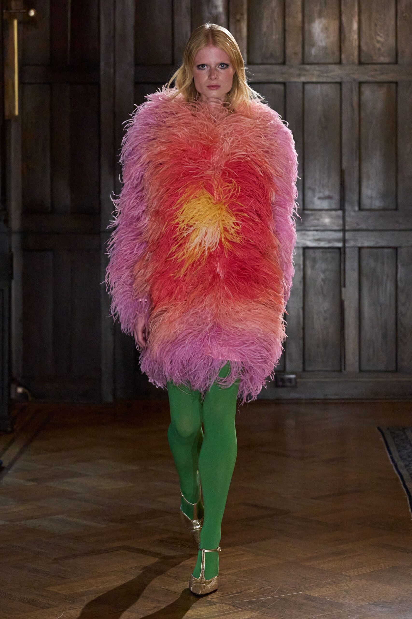Shaggy feather coat in pink-to-coral bands, yellow bursting at the center. Opaque grass-green tights. Gold T-strap heels with a pebbled metallic finish. Walking from a winter wedding into a conservatory full of wet palms.