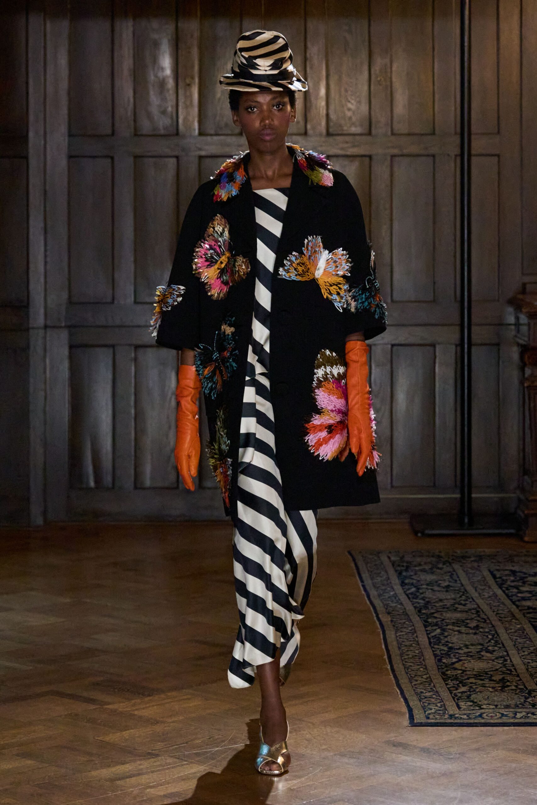 Stacked brimmed hat in cream-and-black zebra stripes. Black swing coat scattered with shaggy multicolour floral appliques. Bias-cut ivory gown banded in wide black diagonal stripes. Burnt orange leather opera gloves. Gold metallic sandals with a pale blue toe strap. Crossing a drafty manor hallway before dinner is called.