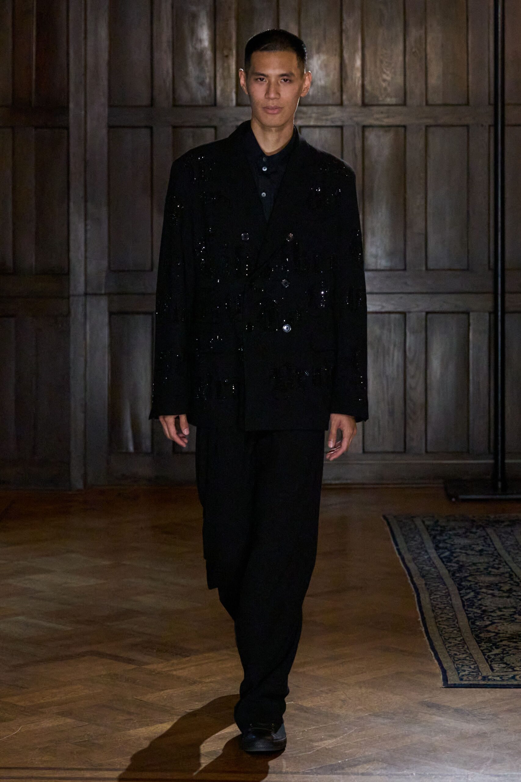 Black double-breasted blazer dusted with sequins and cut with long fringe tiers. Black knit polo, the top button left undone. Loose black trousers falling straight with almost no break. Square-toe black shoes with a dull leather shine. Crossing a wood-panelled hotel hall after the wedding band has packed up.