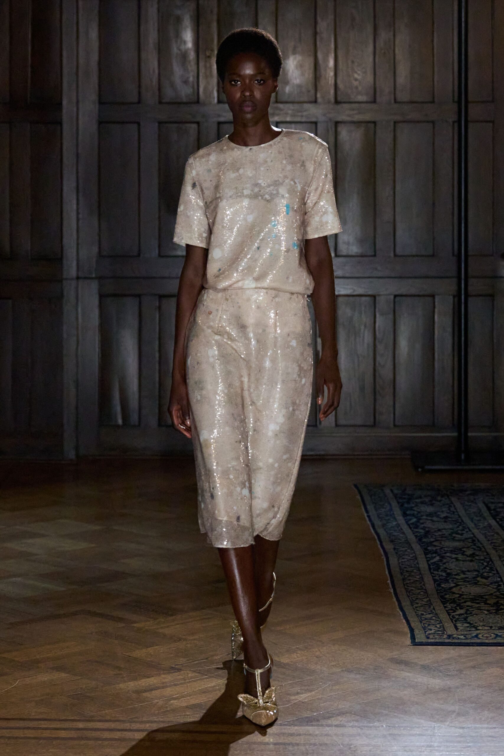Pale beige sequined tee, mottled with cloudy white and turquoise flecks. Matching sequined culottes cut loose through the calf, hem softly rolled. Gold glitter T-strap heels with a butterfly perched on each toe. Crossing a parquet ballroom after the wedding band has packed up.