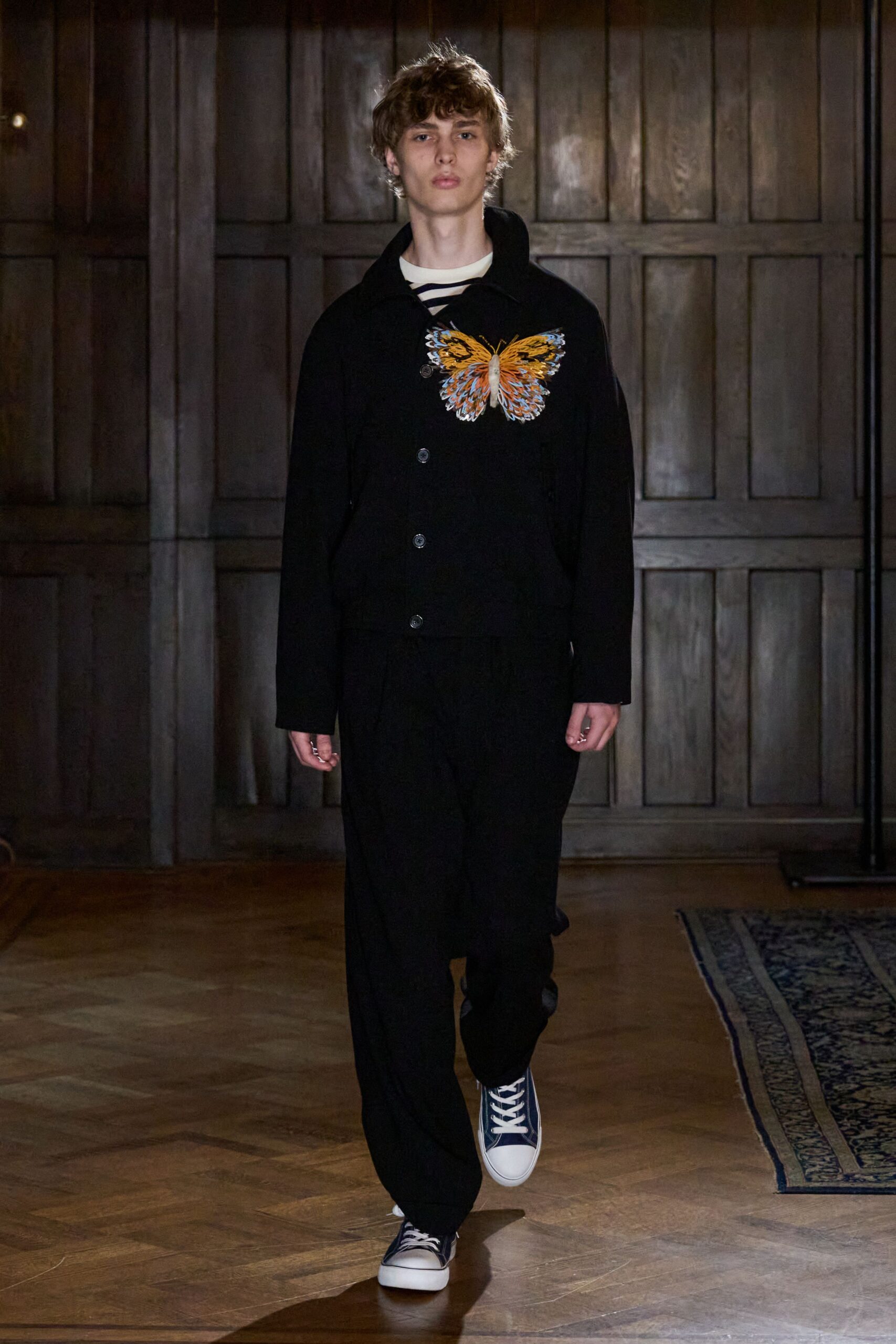 Black wool jacket with a large butterfly appliqued across one chest. Cream crewneck underlayer crossed with navy Breton stripes at the throat. Loose black trousers breaking softly over the shoes. Navy canvas high-tops with white rubber toes. Crossing a drafty manor hallway before anyone has opened the dining room.