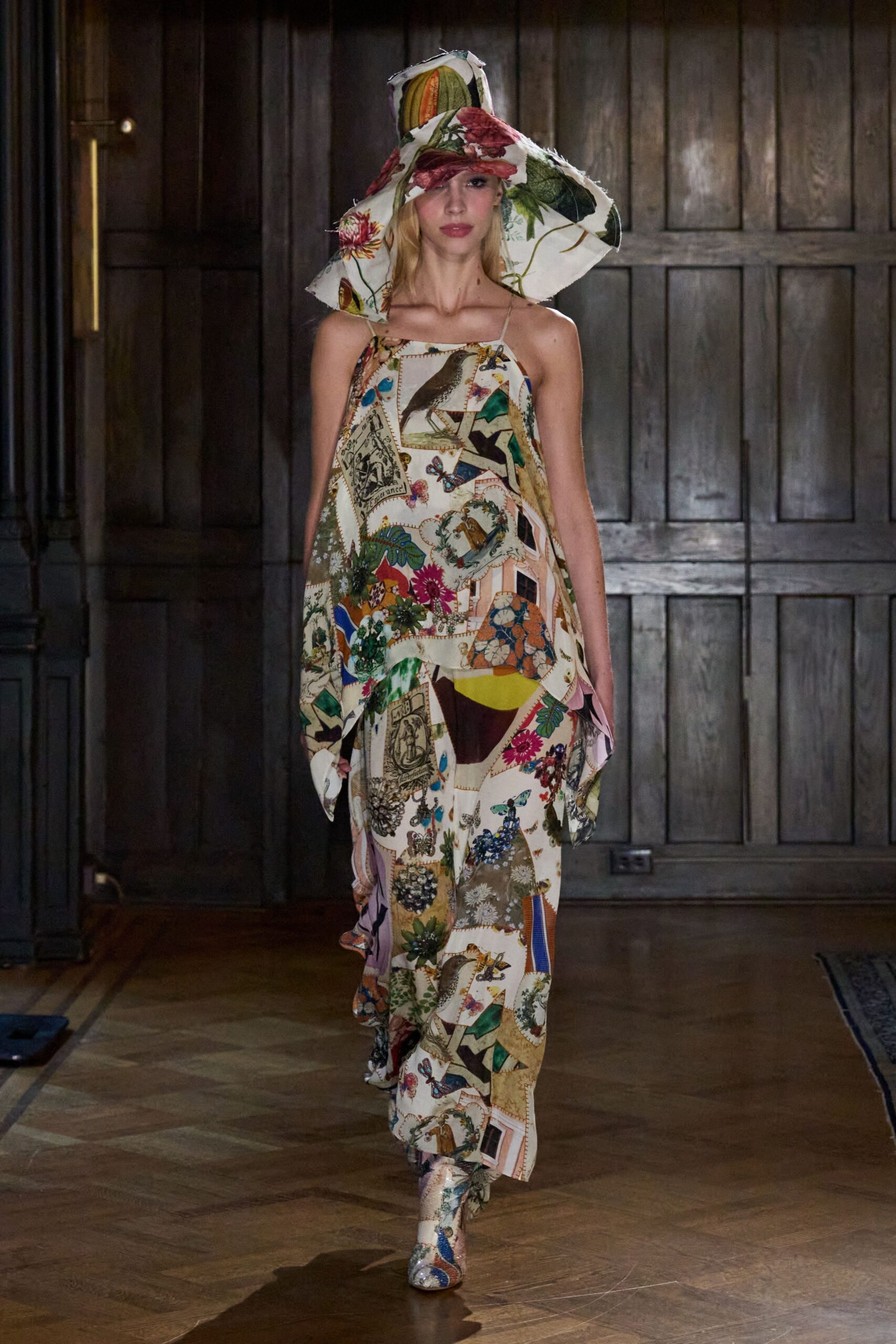 Oversized floppy hat in botanical patchwork, raw-cut brim jutting askew. Handkerchief dress in postcard-and-bird print silk, held by spaghetti straps. Matching printed boots disappearing under the uneven hem. Walking through a conservatory after rain, damp gravel still clinging to the soles.