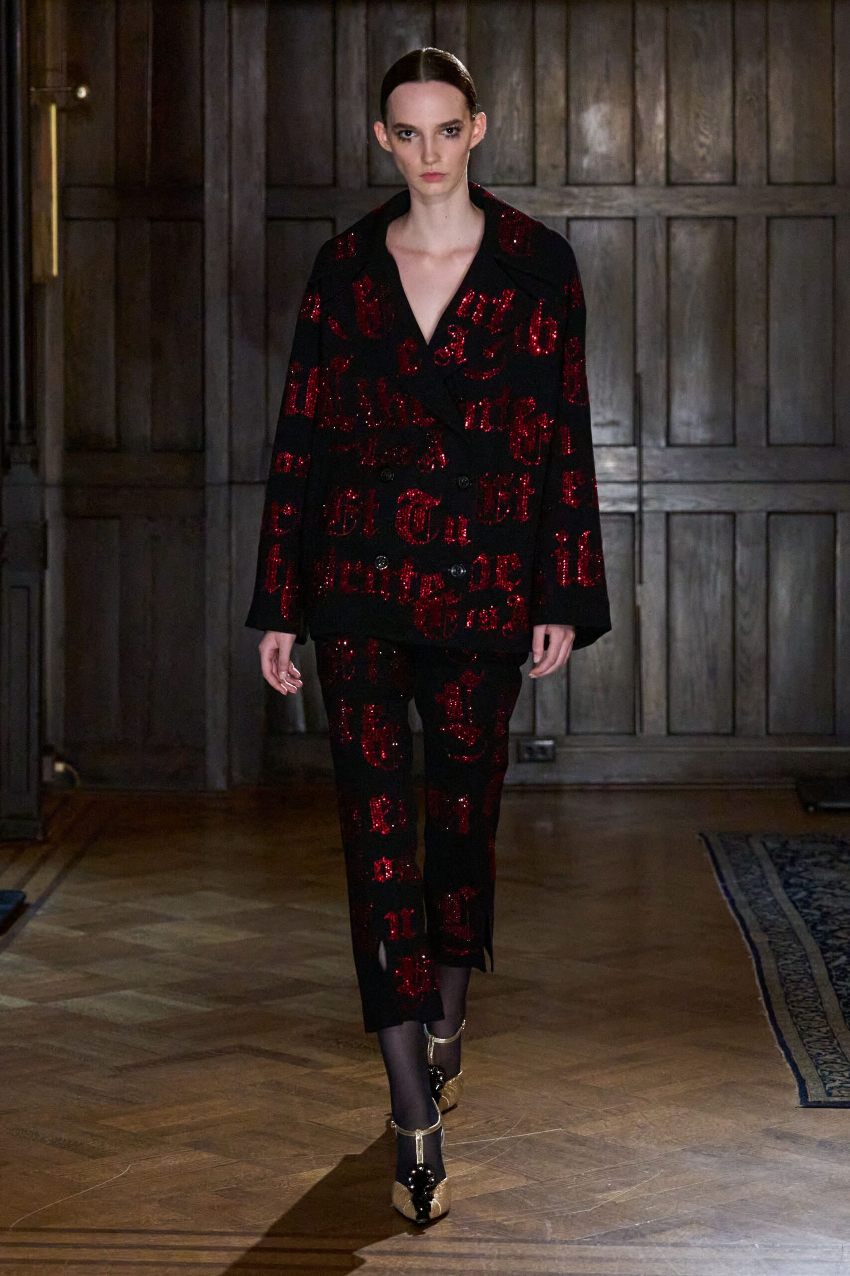 Black double-breasted blazer strewn with crimson sequined Gothic lettering. Slim black trousers matching the sequined script, slit at one hem. Sheer black tights. Gold ankle-strap heels with a black patent cap. Crossing a paneled hotel corridor after the wedding has soured.