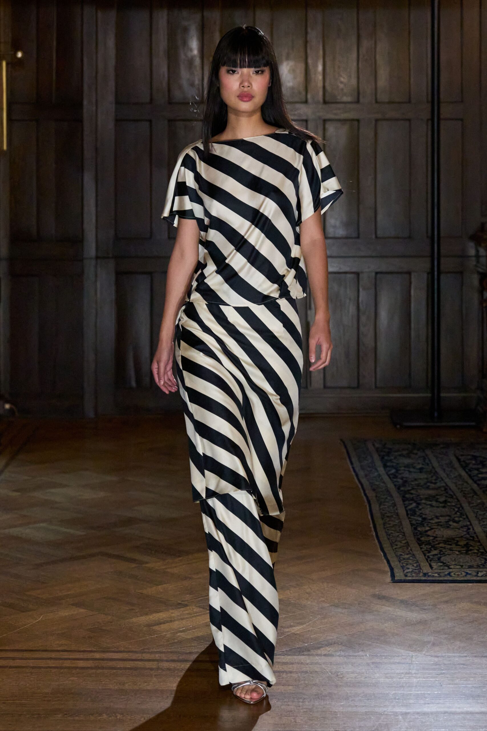 Cream satin top cut on the bias with broad black diagonal stripes. Matching draped satin skirt, ruched at the hip and wrapped over one leg. Clear barely-there straps over a pointed metallic sole. Crossing a panelled hotel lobby before anyone else is downstairs.