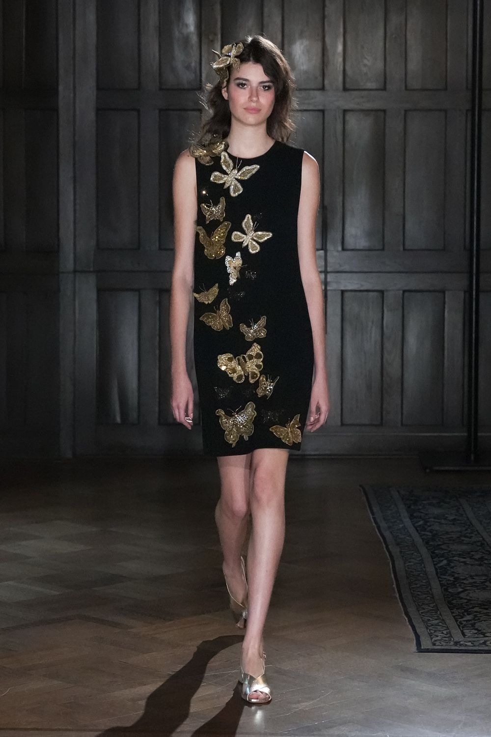 Gold butterfly clip perched above one temple. Black sleeveless shift scattered with raised gold sequined butterflies. Metallic gold peep-toe heels with a narrow slingback. Crossing a parquet ballroom before the string quartet packs up.