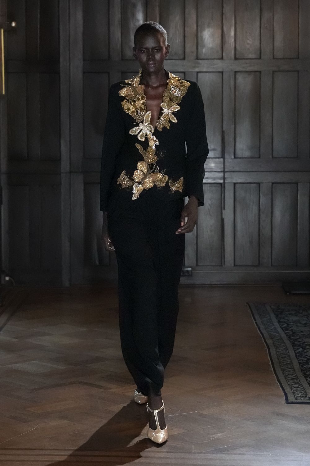 Black crepe top overtaken by dense gold appliqued moths and leaves. Loose black trousers falling straight from the hip. Champagne T-strap heels with a rounded cap toe. Crossing a paneled manor hallway after everyone else has gone upstairs.