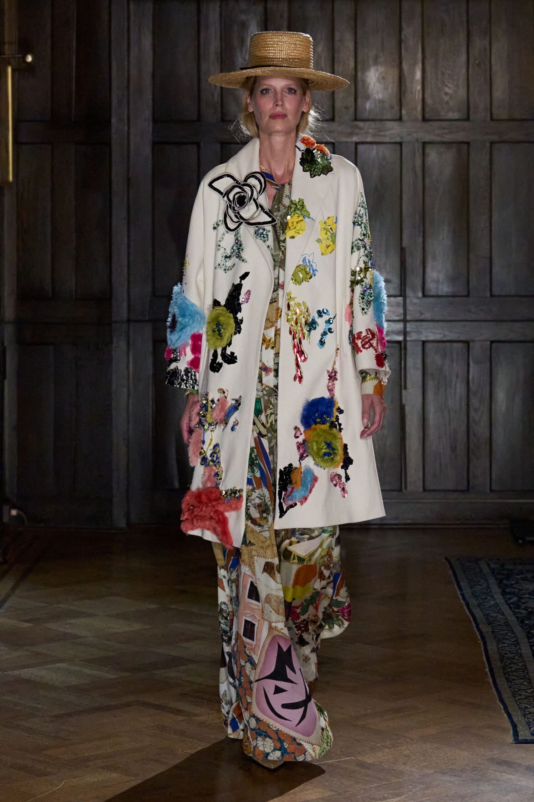 Flat-crowned straw boater with a very wide brim. Ivory coat crowded with sequined blooms and fuzzy pommed appliques. Floor-length collage-print dress, hem pooling over the front foot. Crossing a manor hallway after lunch, still carrying bits of the garden indoors.