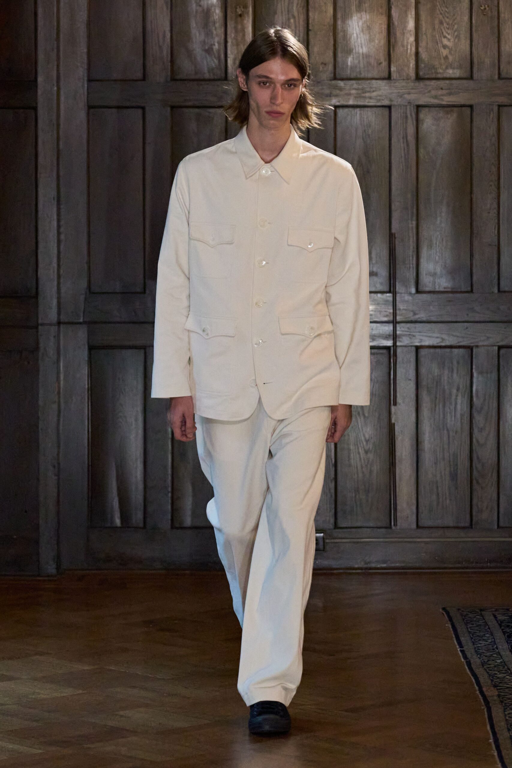 Cream suiting jacket with four scalloped flap pockets and pearly buttons. Matching straight trousers in the same dense cloth, breaking softly at the shoe. Black leather shoes with blunt rounded toes. Crossing a manor hallway before anyone else is awake.