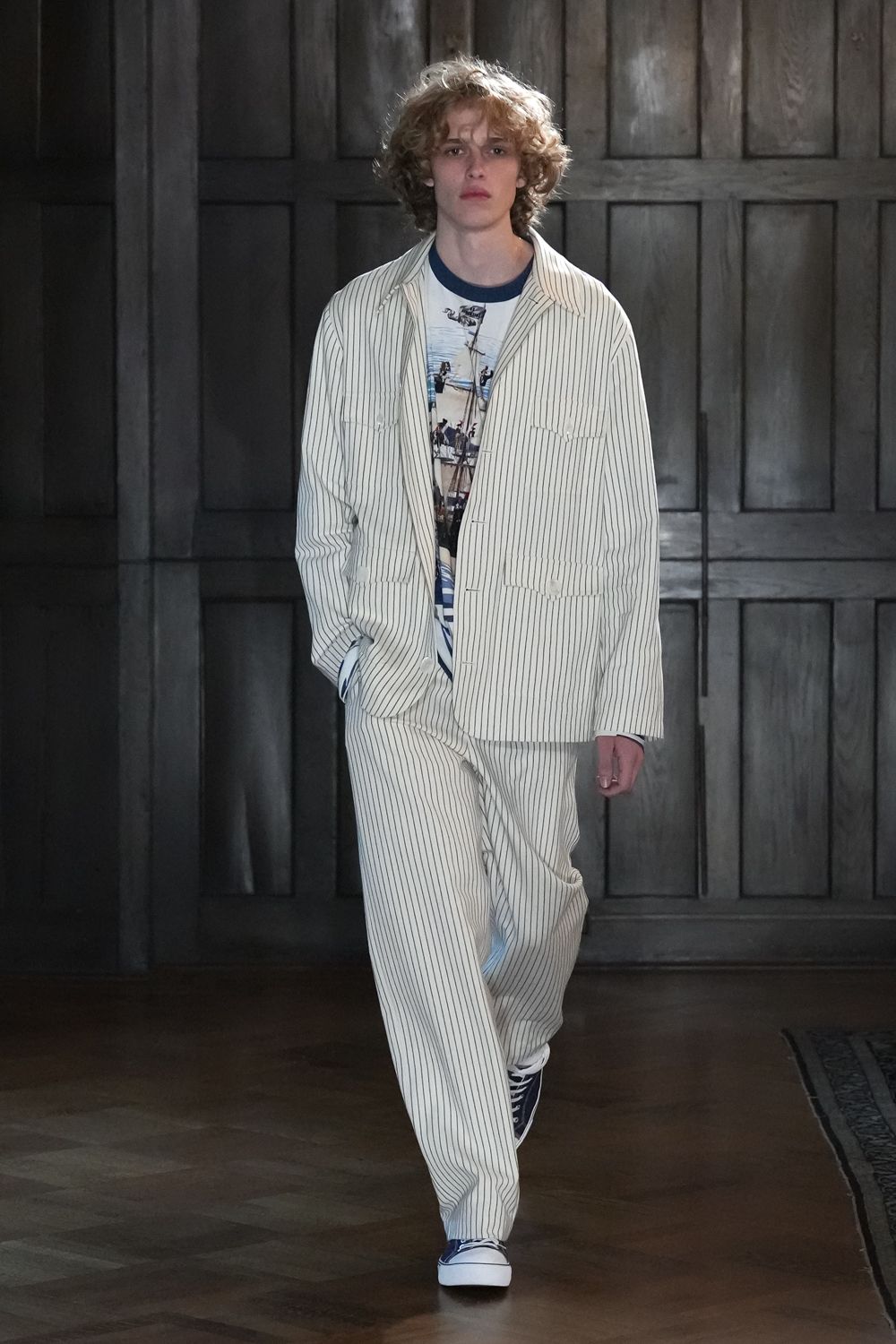 Cream pinstriped work jacket with rounded patch pockets and a soft spread collar. White graphic ringer tee with a navy bound neckline. Matching pinstriped trousers, cut loose and breaking over the sneakers. Navy canvas high-tops with bright white rubber foxing. Walking the long wood corridor before everyone else wakes up.