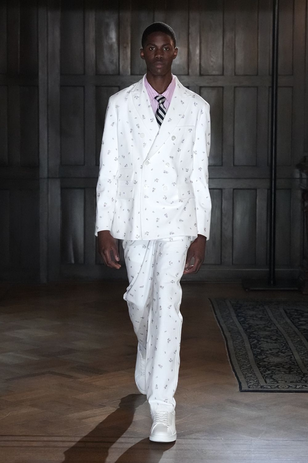 White double-breasted blazer scattered with tiny grey floral sprigs. Pink banker-stripe shirt under the lapels. Black and silver diagonal-striped tie, neatly centered. Matching white trousers, one leg breaking softly over the sneaker. White leather low-top sneakers. For drifting through a paneled house before lunch is announced.