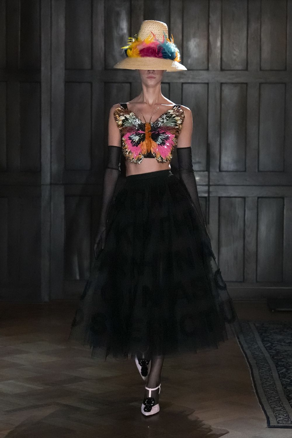 Straw cloche trimmed with a ring of bright feathers. Butterfly-wing bodice in layered sequins, antennae rising at the neckline. Sheer black opera gloves. Black tulle ball skirt, faint circles drifting through the layers. Pale T-strap pumps with black floral rosettes. Garden party after rain, grass still slick under the tables.