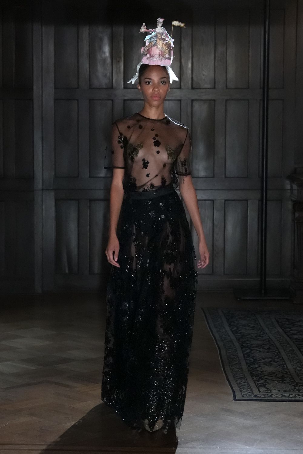 Mini rococo figurine hat with ribbon streamers and a tiny pennant. Sheer black tulle top scattered with sequined floral appliques. Floor-length black mesh skirt densely encrusted, hem ghosting over the shoes. Crossing a drafty manor hallway after midnight cake has gone untouched.