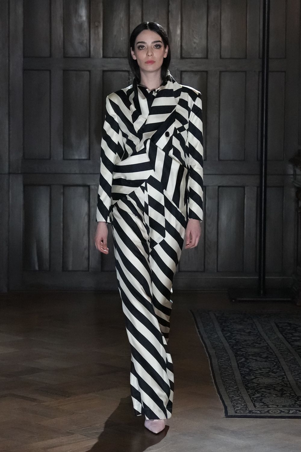 Ivory satin blazer cut double-breasted, black stripes breaking at the lapels. Matching satin trousers with diagonal stripes skewed across the fly. Pale pointed pumps peeking under the hem. Crossing a wood-panelled hotel corridor before the wedding breakfast.