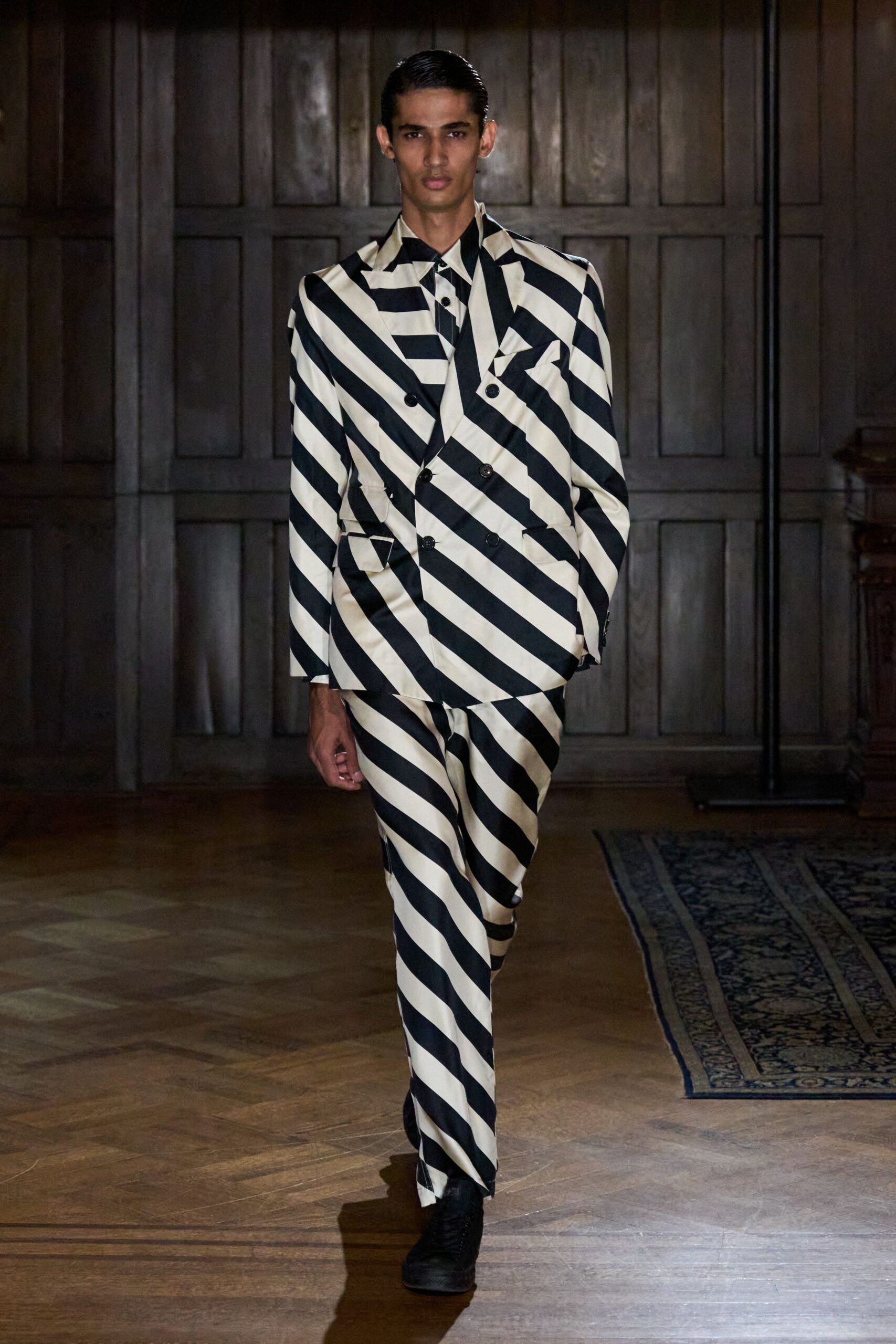 Cream satin double-breasted blazer cut in wide black barber-pole stripes. Matching striped shirt with a small black button at the throat. Straight satin trousers, stripes swinging diagonally into the break. Black lace-up shoes. Crossing a parquet hall before the string quartet has finished tuning.