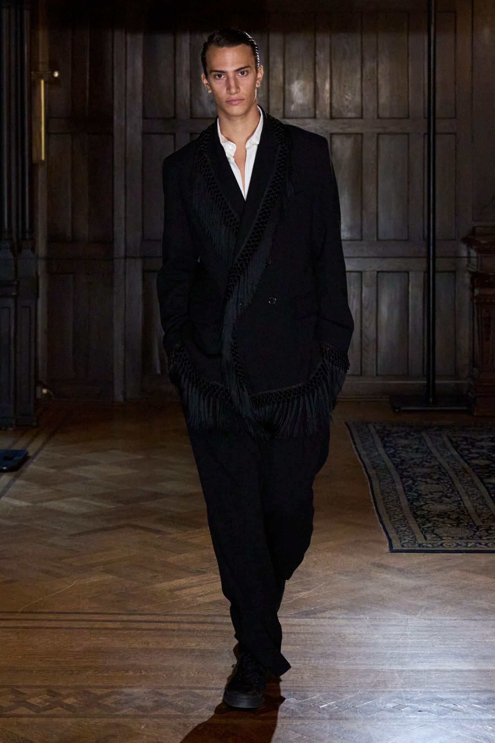 White shirt worn open at the throat, collar slightly rumpled. Black double-breasted blazer traced with braided trim and long fringe. Black straight trousers breaking softly over the shoe. Black low-top sneakers, matte and nearly swallowed by shadow. Crossing a paneled hotel lobby after midnight, room key already in hand.