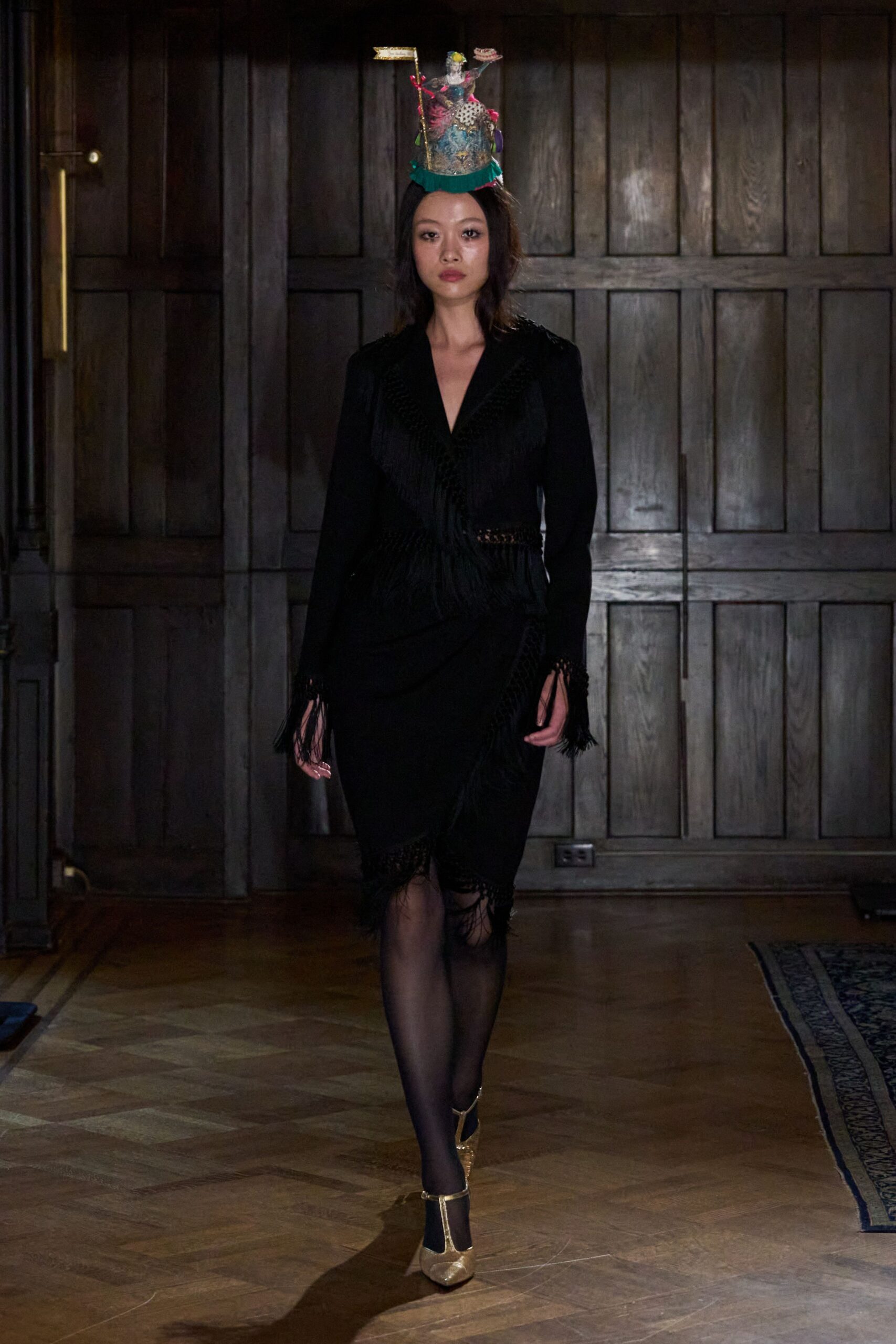 Miniature cake hat with piped icing, tiny topper and green ruffle brim. Black wrap dress cut with ladder-knit waist insets and long fringe. Sheer black tights. Gold T-bar heels, the pointed toes catching the floorlight. Crossing a paneled ballroom after the birthday cake has already been cut.