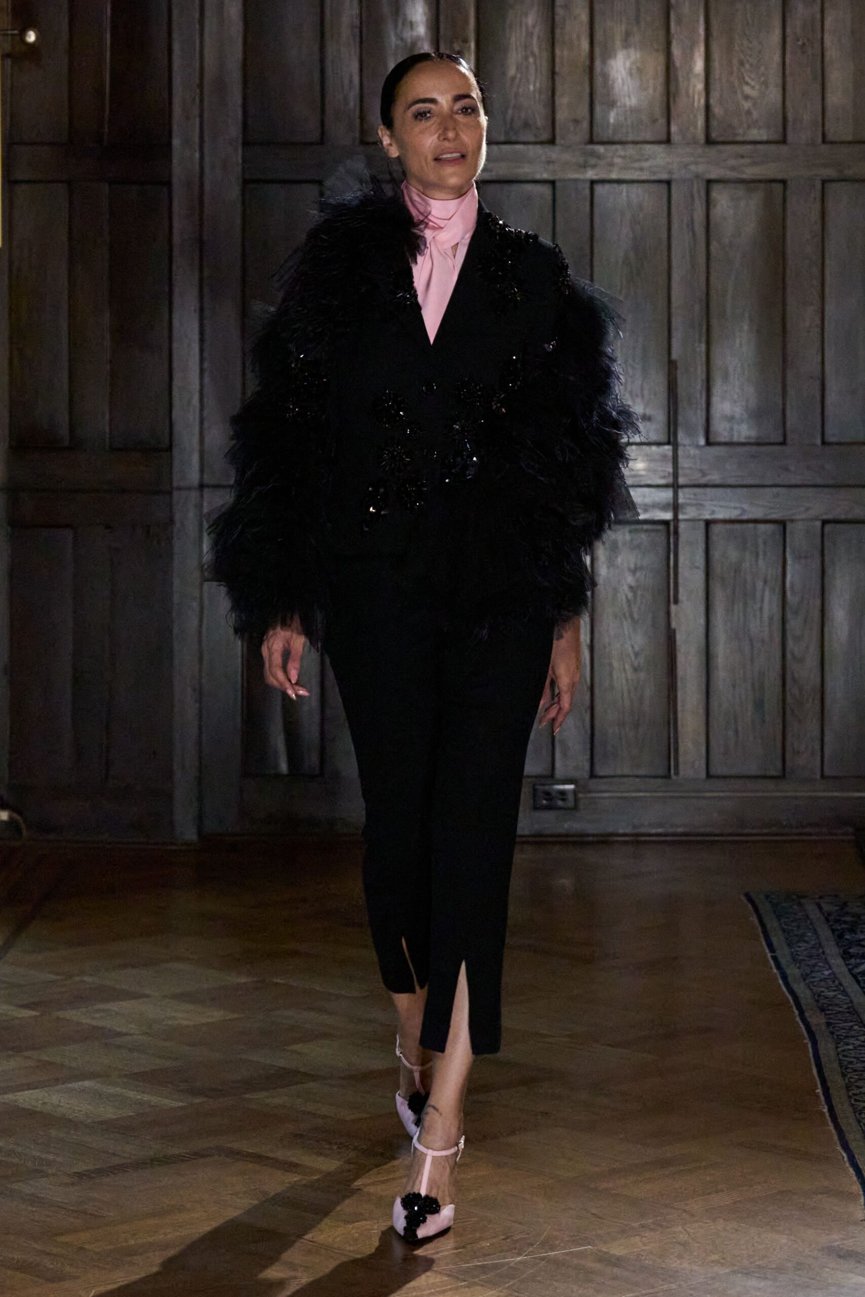 Pale pink blouse with a narrow scarf neck tucked high. Black jacket engulfed in shaggy feathered sleeves, sequins glinting at the waist. Slim black trousers with a neat slit breaking at the front hem. Blush T-strap pumps topped with a black beaded flower. Crossing a parquet ballroom before anyone has turned the chandeliers on.