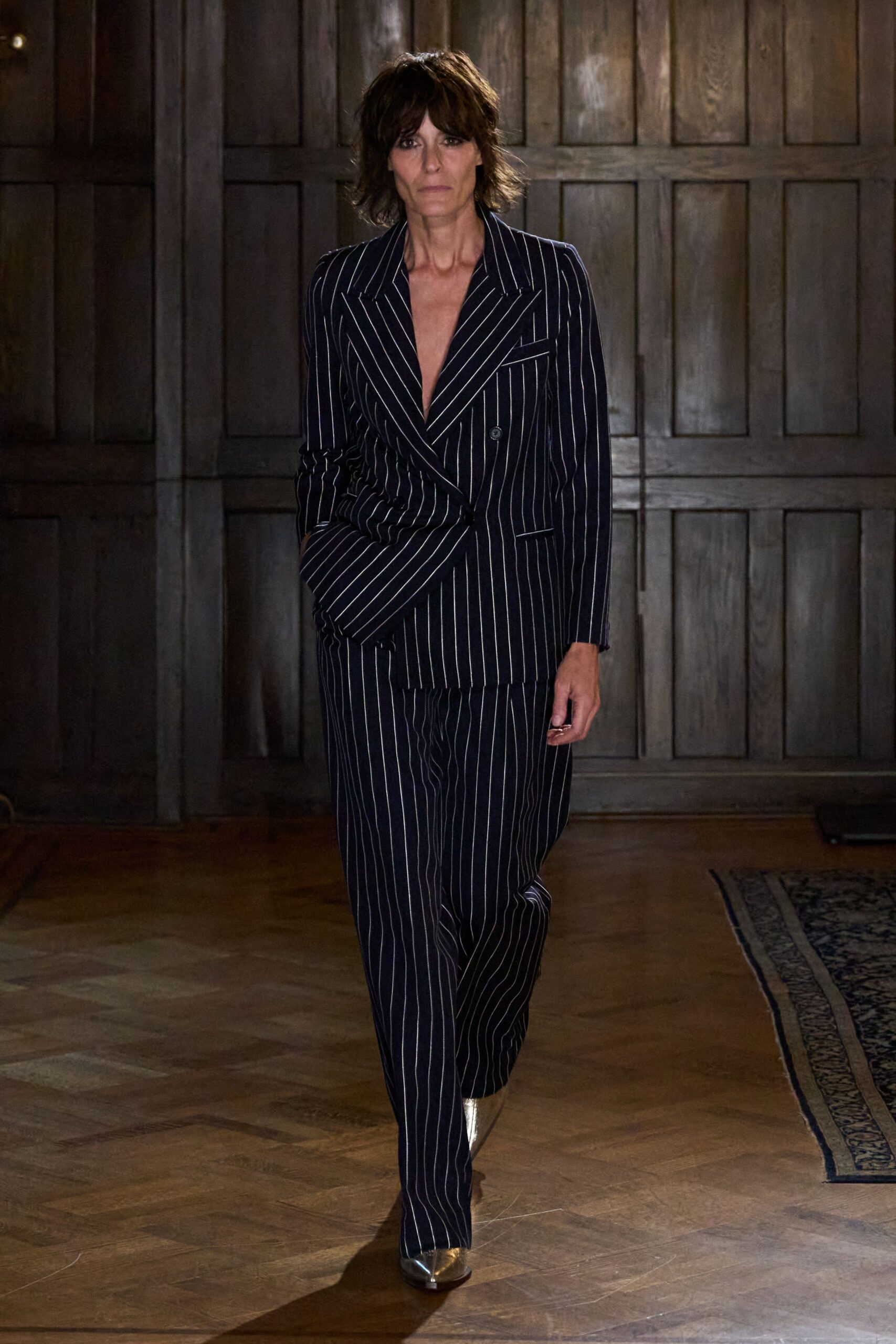 Ink pinstripe double-breasted blazer cut to a bare plunging V. Matching pinstripe trousers falling straight and long over the shoe. Silver pointed boots flashing from the hem. Crossing a parquet hall after midnight, still carrying the speech in your head.