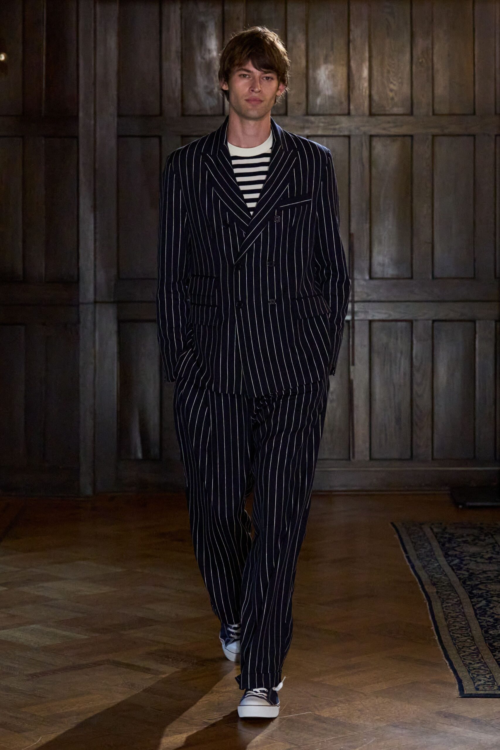 Navy double-breasted blazer cut in chalk pinstripes, one low button fastened. Cream tee banded with navy Breton stripes. Matching pinstriped trousers with a loose straight fall. Navy canvas sneakers, white foxing bright at the toe. Crossing a wood-panelled hotel lobby before breakfast service starts.