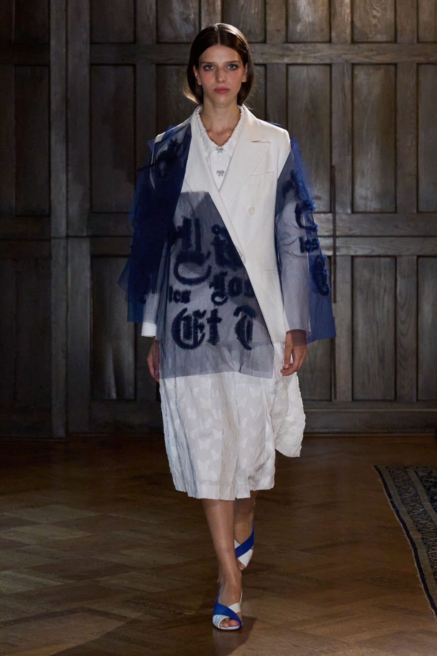 Navy tulle overlay printed with blurred blackletter across one shoulder. White double-breasted blazer, one side left bare beneath the tulle. White jacquard dress with a puckered hem and tiny jeweled closures. White sandals crossed with cobalt bands. Crossing a wood-panelled hall after the invitation said formal but not obedient.