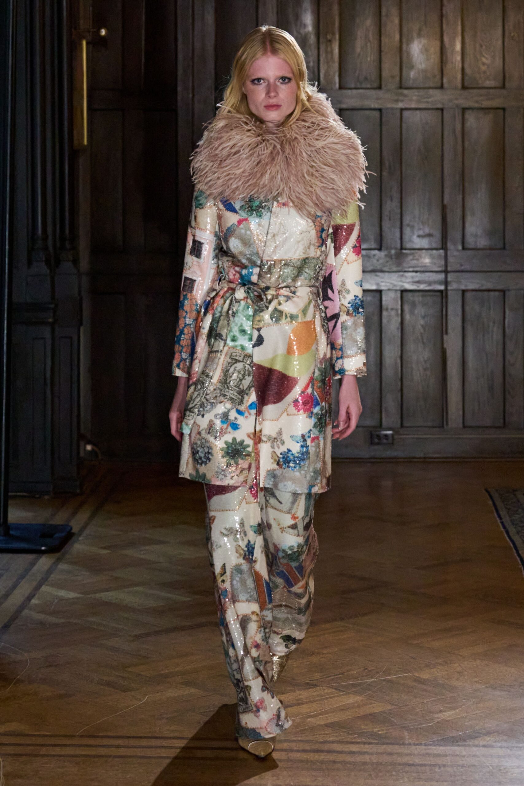 Blush ostrich-feather stole ballooning high around the neck. Cream sequined coat printed like a scrapbook, self-belt knotted off-center. Matching wide trousers pooling over the front of the shoe. Pointed gold boots flashing beneath the hem. Crossing a drafty manor hallway before dinner is announced.