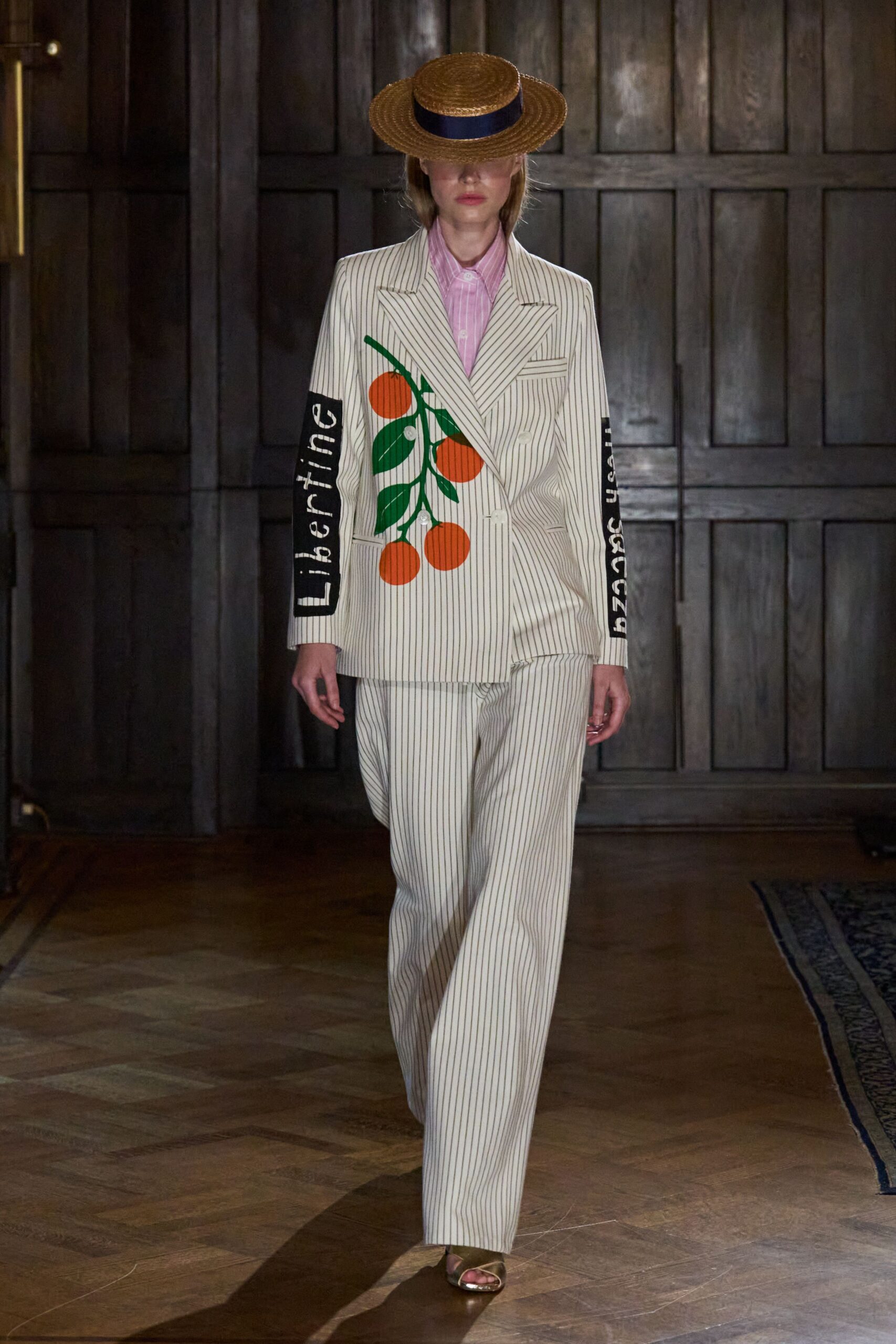 Flat straw boater with a navy grosgrain band. Cream pinstriped double-breasted blazer, one side printed with hanging oranges. Pink striped button-up, collar points tucked under the lapels. Matching pinstriped trousers cut loose through the leg. Olive satin sandals. Crossing a citrus estate dining room before lunch is called.