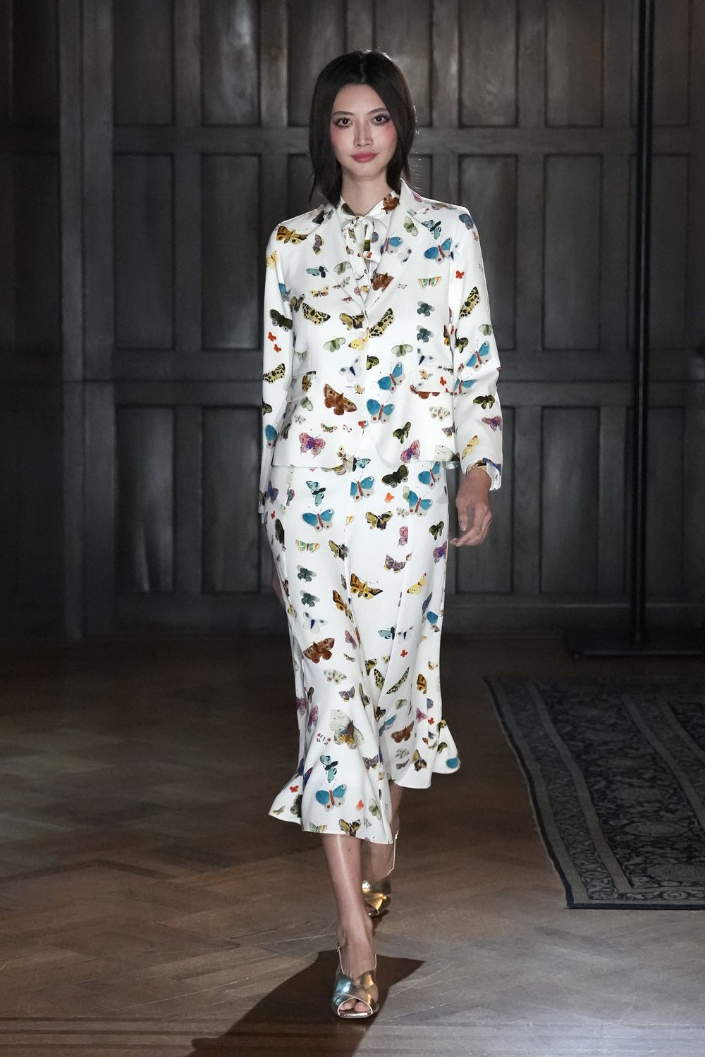 Ivory silk blouse scattered with naturalist butterflies, neck tied in a soft bow. Matching butterfly-print trousers, cut long and slightly fluted at the hem. Mirror-gold peep-toe heels. Crossing a manor hallway before lunch is called from another room.