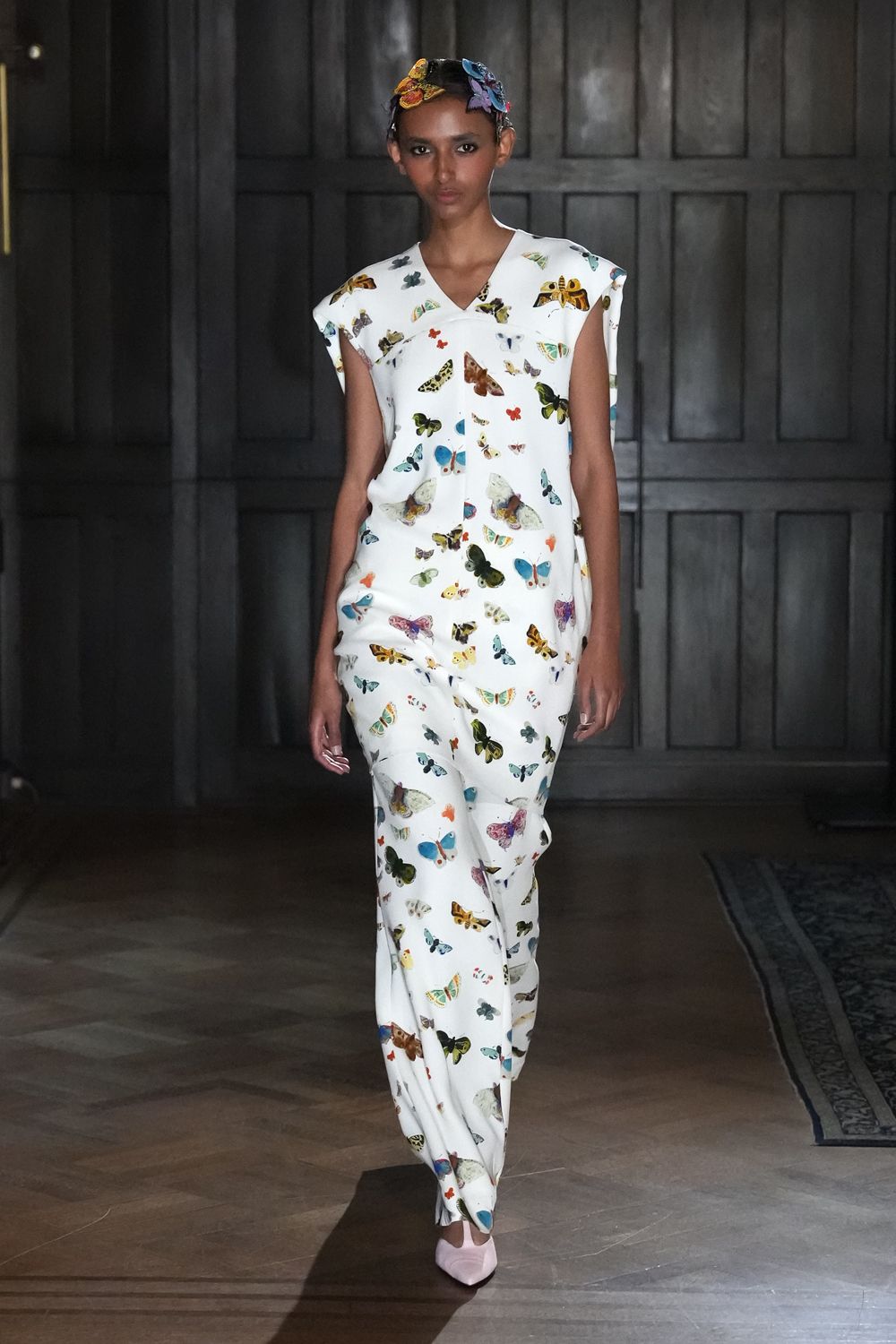 Butterfly appliques pinned across the hairline, one bright blue at the temple. White column dress printed with moths and butterflies, shoulder flanges jutting outward. Pale pink pointed pumps. Crossing a drafty manor hallway after opening the cases in the insect room.