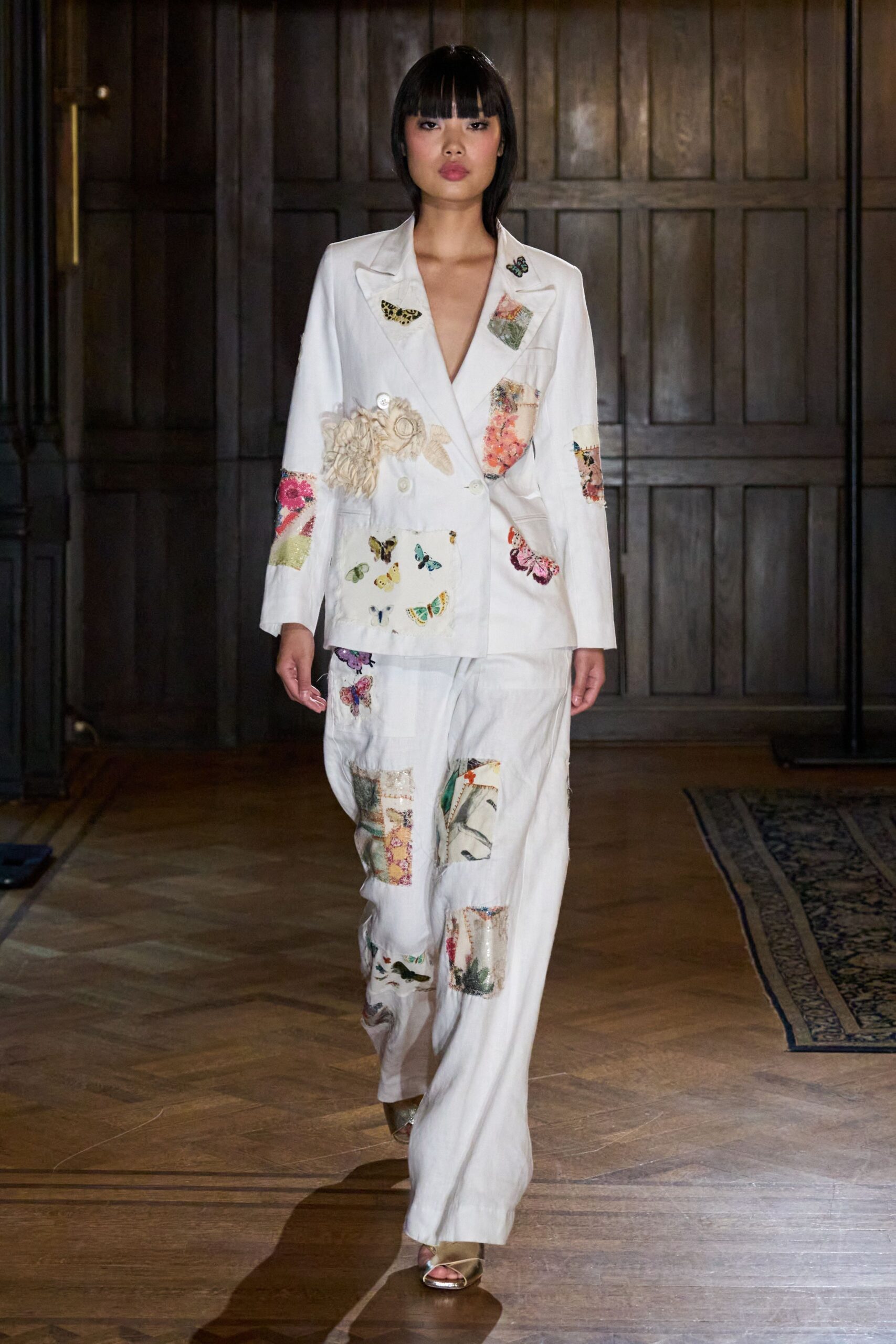White double-breasted blazer patched with butterflies, florals and a rosette at the waist. Loose white trousers scattered with mismatched appliqué squares and a bird at the cuff. Gold cross-strap sandals. Crossing a creaky manor hallway after raiding the sewing room.