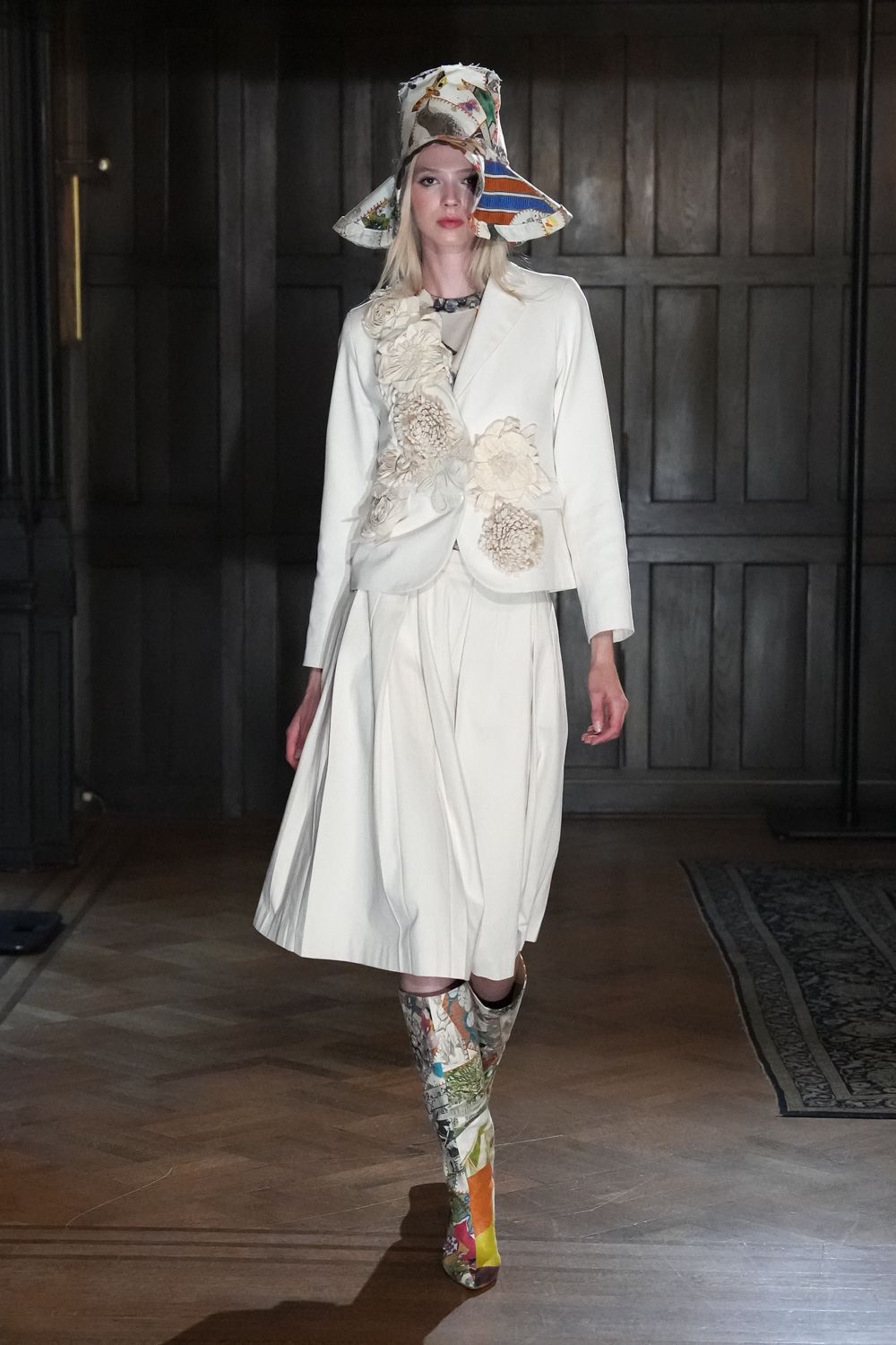 Folded paper-print hat with two wide corners jutting off the head. Ivory tailored jacket smothered in oversized ruched fabric flowers. Soft white dress falling in deep gathered folds below the jacket. Knee-high boots patched from maplike scenic print panels. For reading a will in a country house after the rain stops.
