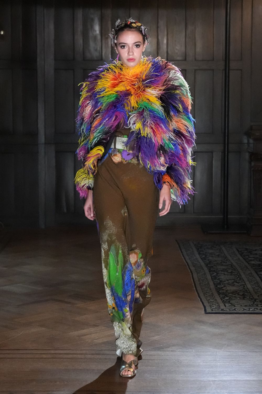 Silver foil leaves and butterfly scraps wired into the hair. Rainbow feather jacket blown huge at the shoulders and throat. Brown draped trousers airbrushed with painterly faces and acid green swathes. Metallic platform sandals with clear block heels. Crossing a drafty manor hallway after midnight, still wearing the craft table.