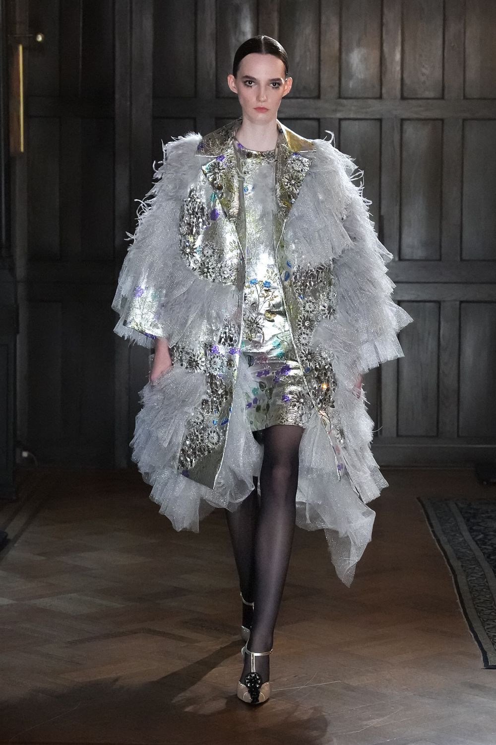 Silver brocade coat buried under tiered ice-grey tulle ruffles. Matching metallic mini with foil shine and scattered violet flashes. Sheer black tights. Champagne T-strap heels with a black rosette clipped at the toe. Crossing a draughty manor hallway after midnight champagne.