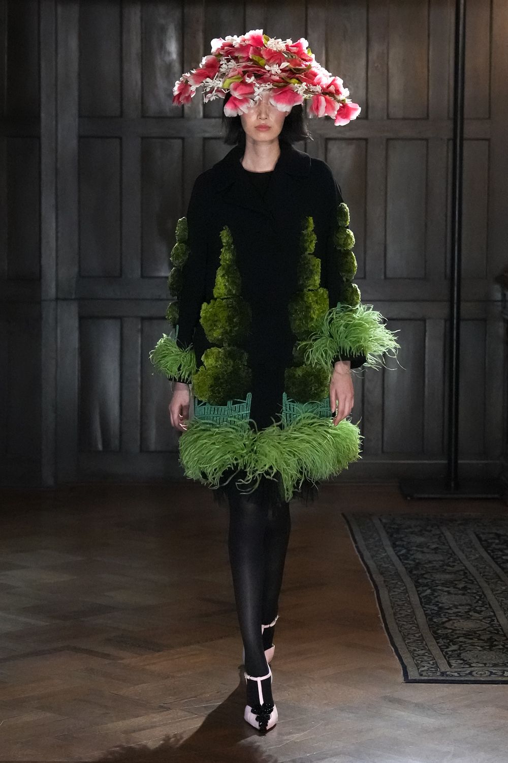 Oversized brim heaped with pink-and-white silk blossoms over the eyes. Black coatdress appliqued with clipped topiary towers and mint ostrich cuffs. Opaque black tights. Blush ankle-strap heels with a black beaded flourish at the toe. Crossing a manor hallway after raiding the conservatory for centerpieces.
