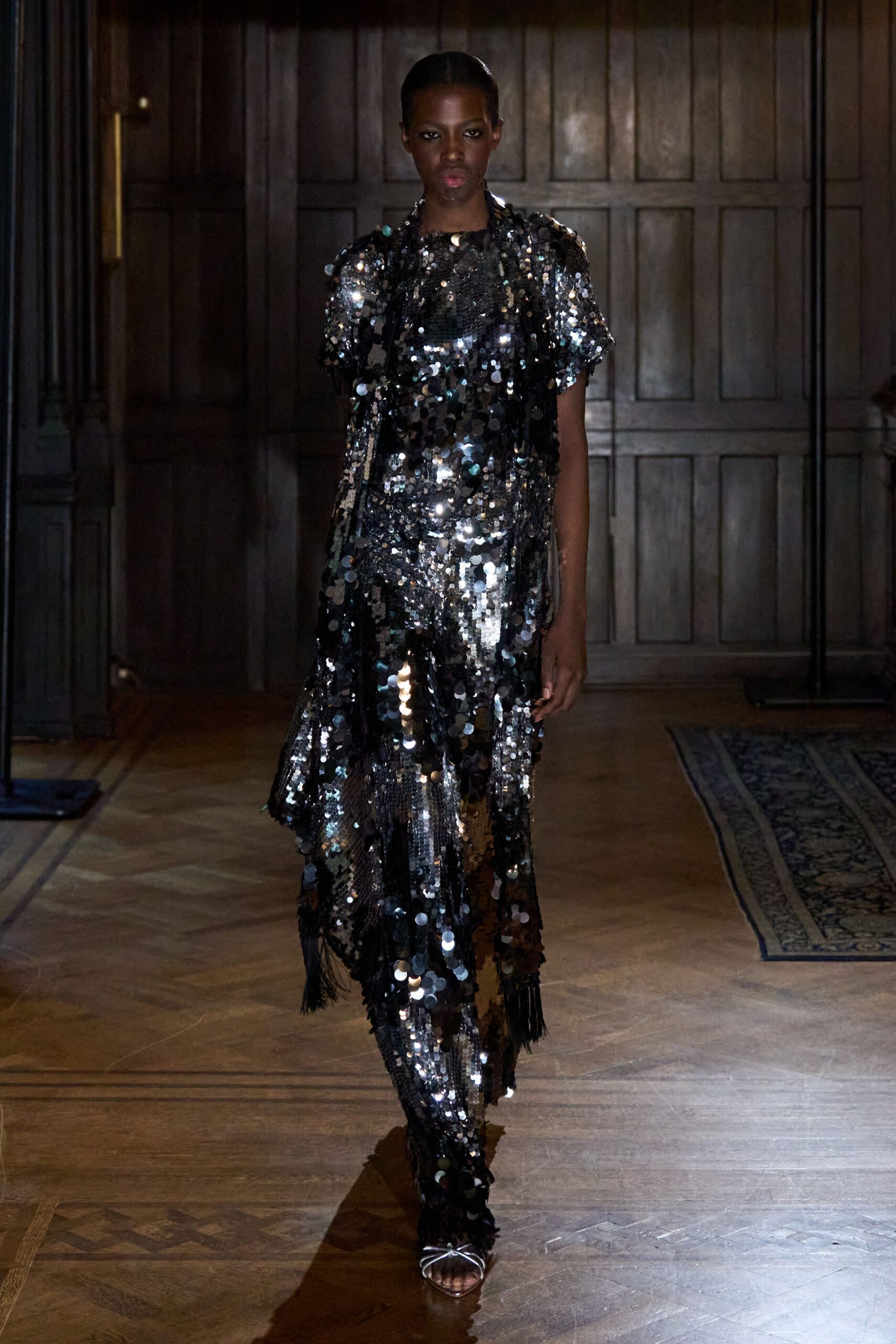 Black sequin top with chunky paillettes and short rounded sleeves. Long black fringe scarf draped loose, one end trailing past the knee. Matching sequin trousers, mirror discs catching light in vertical streaks. Silver thong sandals. Crossing a paneled hotel bar after the quartet has packed up.