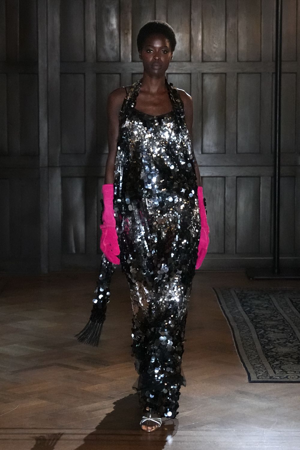 Loose silver sequin column gown with oversized paillettes and a low square neck. Hot-pink gloves in plush suede. Black beaded fringe pouch hanging long from one hand. Silver barely-there sandals peeking beneath the hem. Crossing a parquet ballroom after midnight, still carrying the party in one hand.