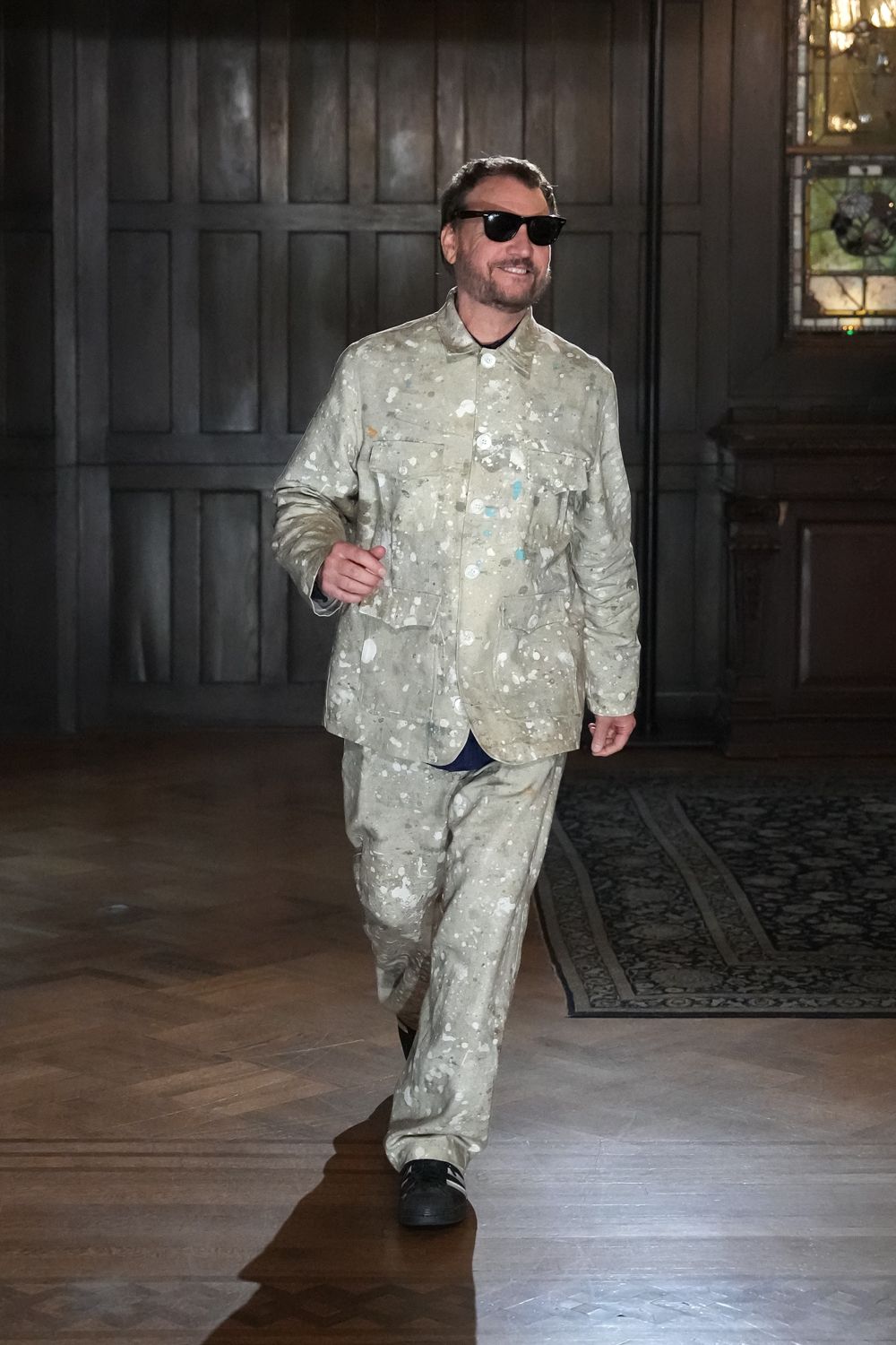 Black cat-eye sunglasses. Pale sage shirt jacket speckled all over with paint-spatter marks. Matching loose trousers, hems breaking softly over the shoes. Black sneakers with white side stripes. Crossing the manor hallway after hanging the last canvases.