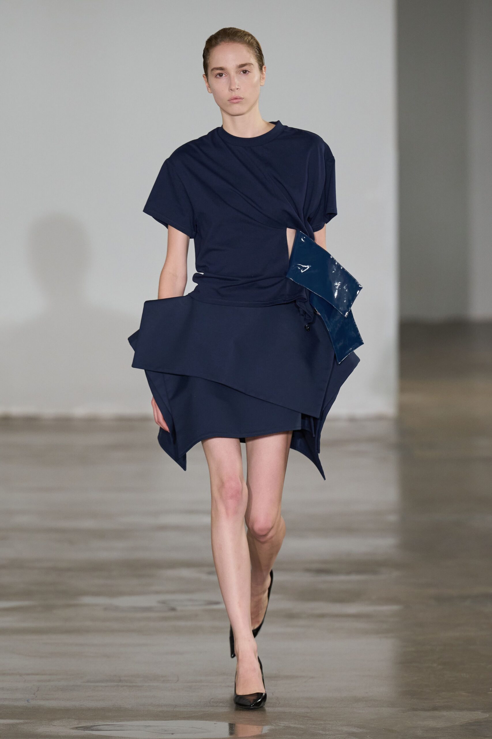 Navy jersey top twisted across the torso with a sharp side cutout. Asymmetric navy mini skirt in stiff suiting cloth, folded into jutting planes. Glossy petrol patent flap hanging from the waist, one corner kicked outward. Black patent pumps. For crossing a museum atrium when the floor still smells faintly of rain.