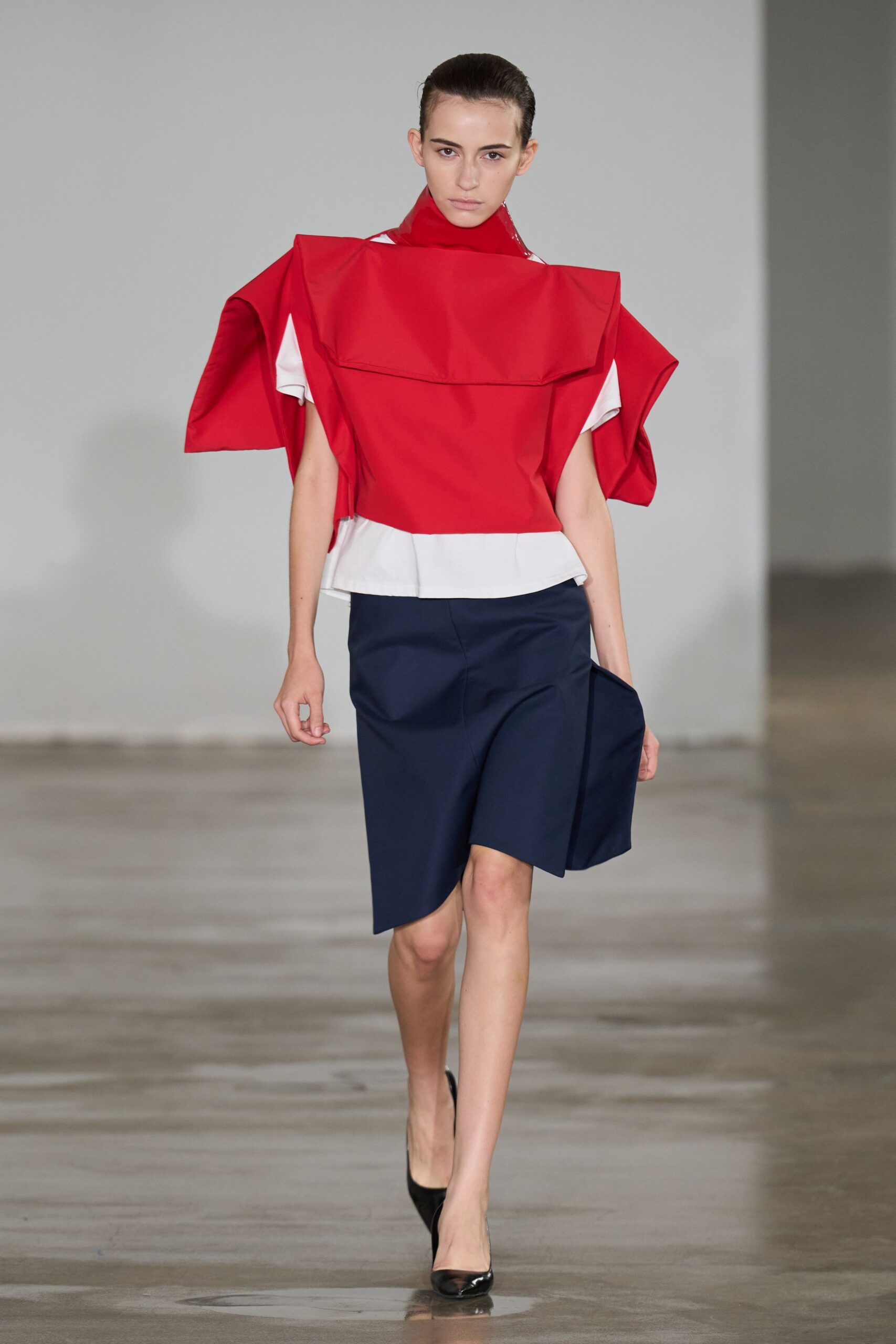 Red cotton shell with exaggerated folded shoulder wings and a bib-like flap. White high-neck insert peeking at the sleeves and kicked peplum hem. Navy wrap skirt cut on a sharp diagonal, one panel jutting forward. Black patent pointed pumps. Walking into a municipal atrium before the architecture tour starts.