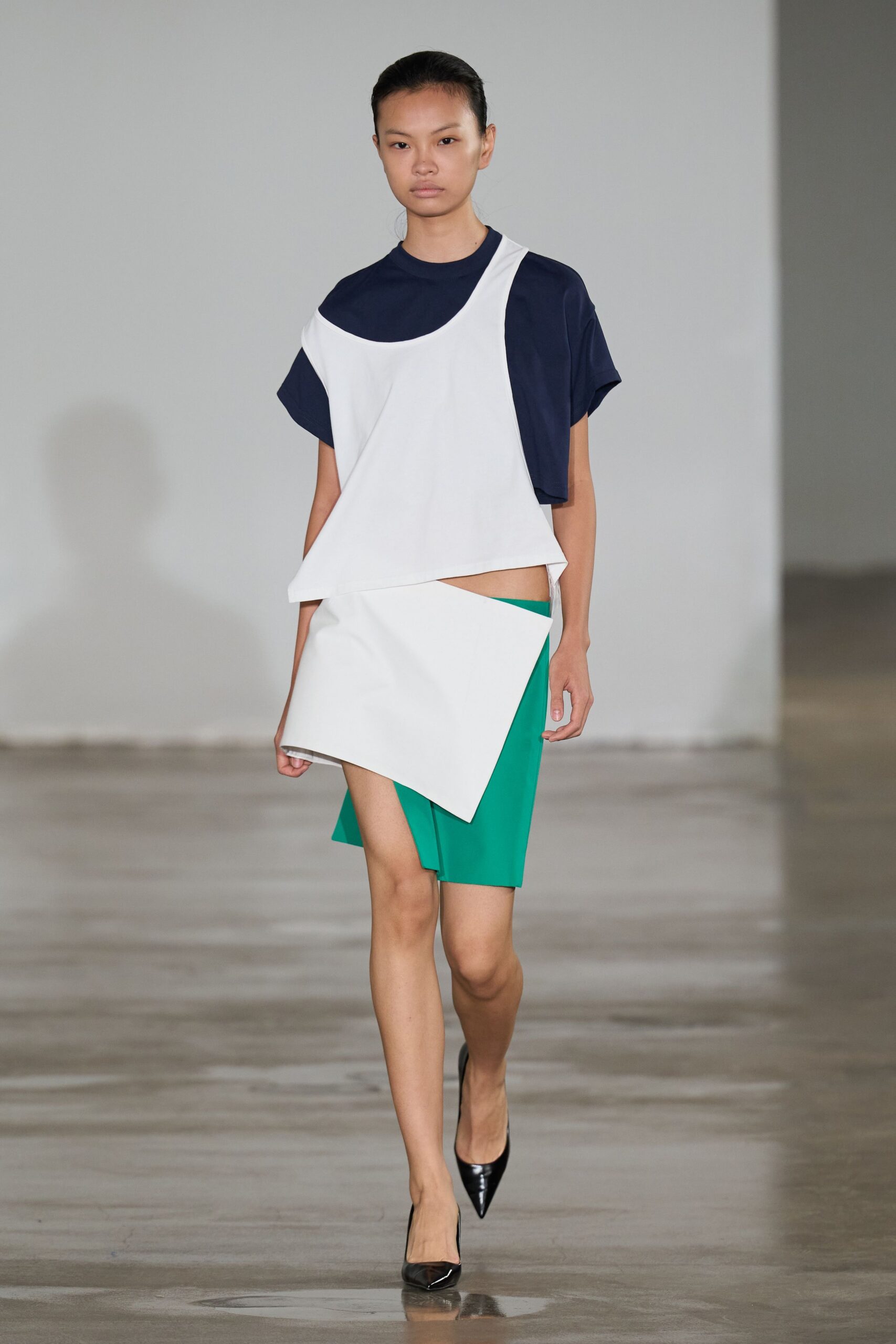 Navy jersey tee with a dropped shoulder and close crew neck. White bonded panel wrapping diagonally, one side hanging past the waist. Emerald skirt cut on a sharp slant, bare hip peeking through. Black patent pumps with a narrow pointed toe. Crossing an empty museum atrium before the guards unlock the side doors.