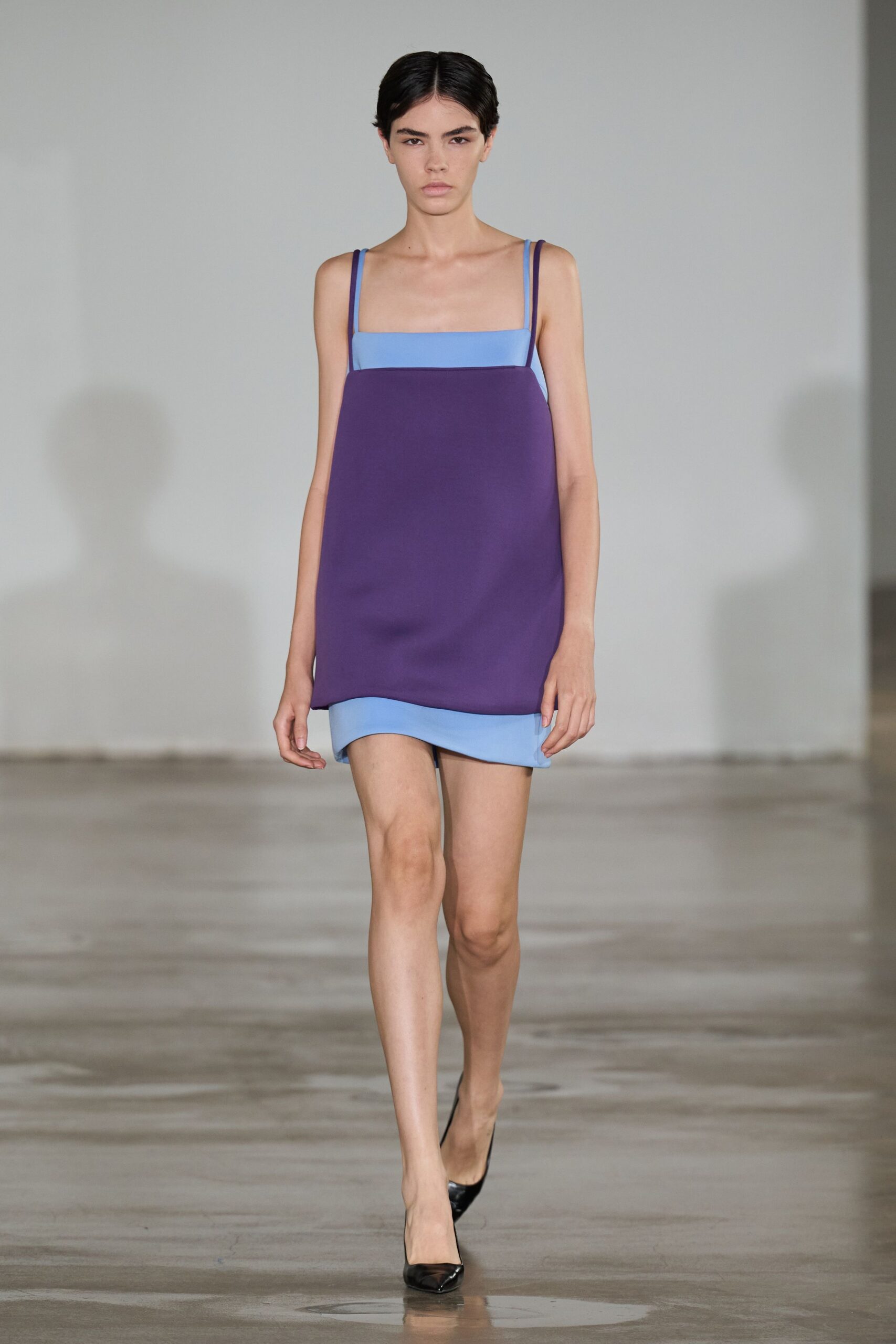 Powder-blue mini slip with a square neckline and narrow straps. Deep violet tunic layer hanging straight, hem kicked up at one corner. Black patent pumps with a sharp, pinched toe. Early check-in at a concrete hotel, suitcase still not unpacked.