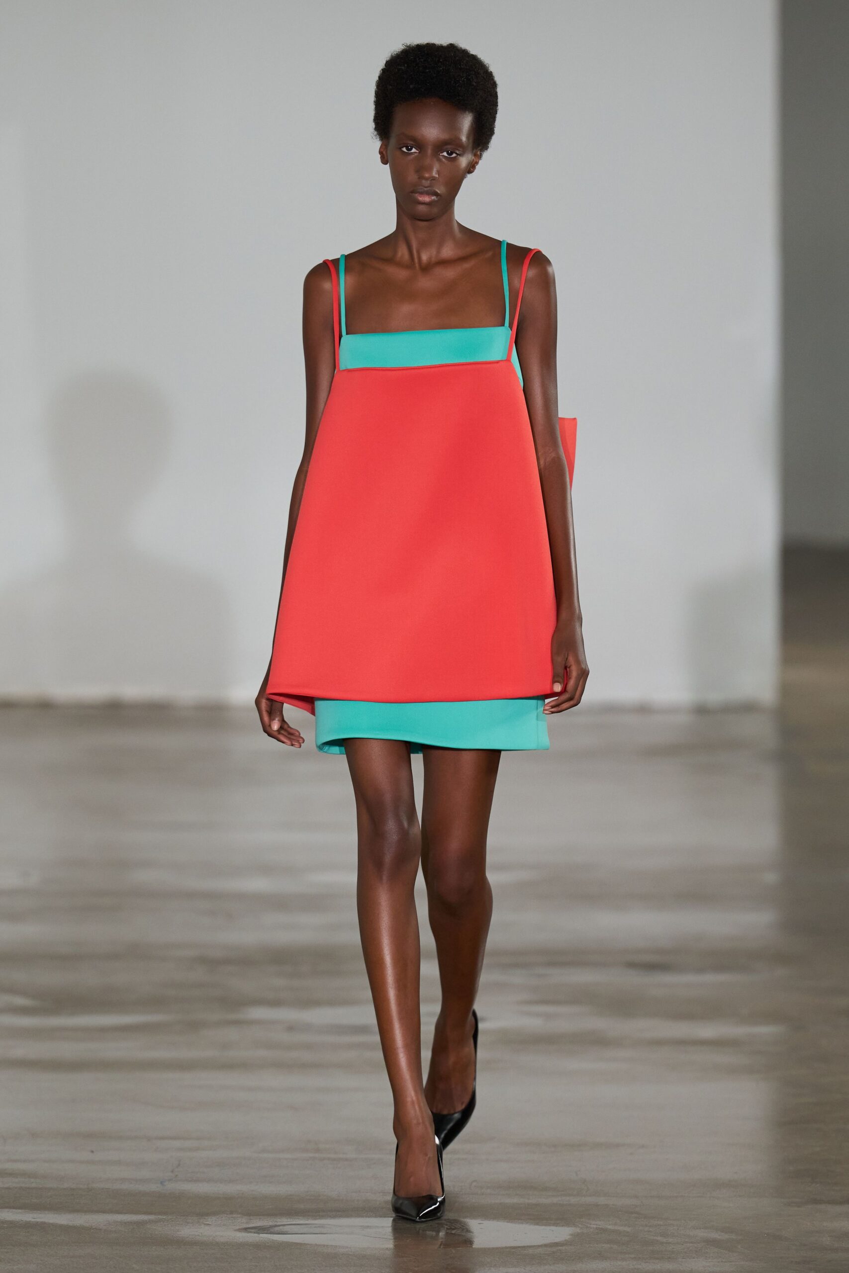 Boxy coral mini in stiff bonded cloth, turquoise underlayer peeking below. Mismatched slim shoulder straps in coral and turquoise. Black patent pumps with a sharp pointed toe. Walking through a design fair with rolled posters under one arm.