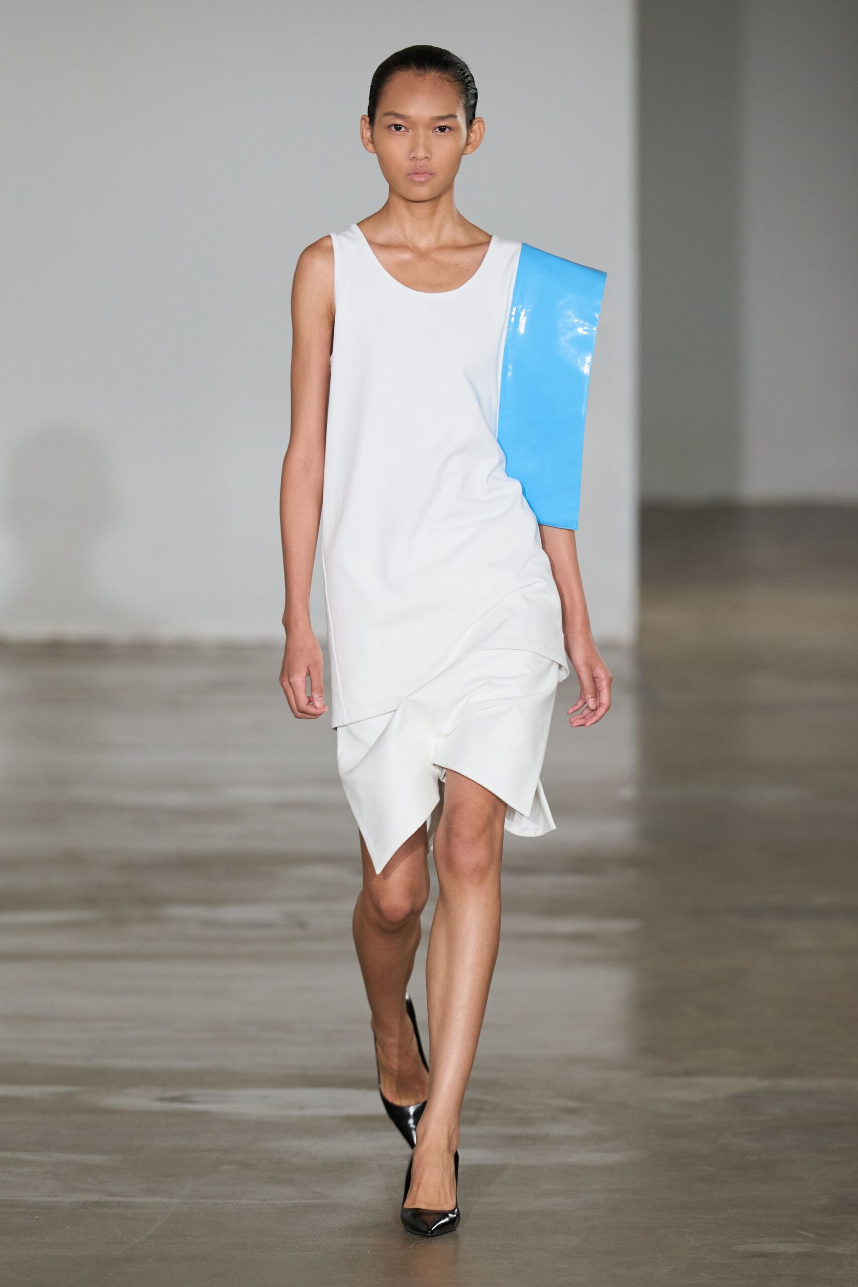 White draped shift dress with an angular hem folding over one knee. Glossy sky-blue vinyl slab dropping from one shoulder like a half sleeve. Black patent pumps, sharply pointed. Crossing a near-empty atrium after the rain finally stopped.