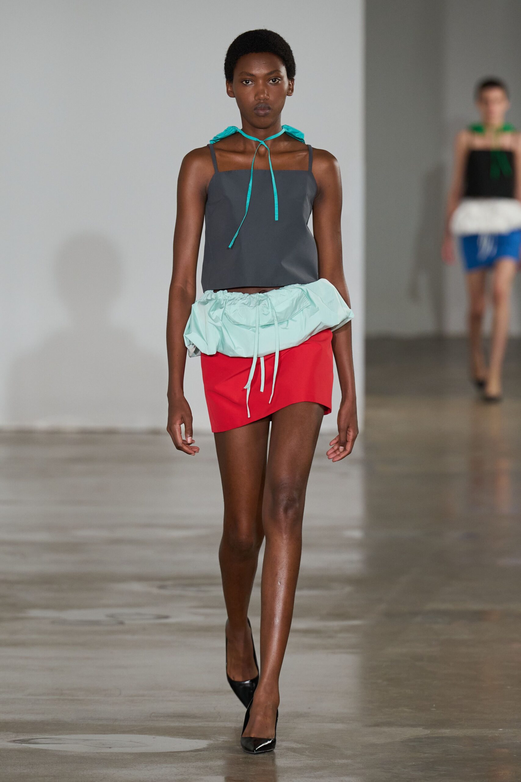 Charcoal camisole with a straight neckline and narrow shoulder straps. Turquoise nylon ties looped at the throat, ends hanging unevenly. Red mini wrapped with a puffed mint drawstring tunnel sitting low. Black patent pumps. Walking the pier after rain, wind still trapped inside the anorak hem.