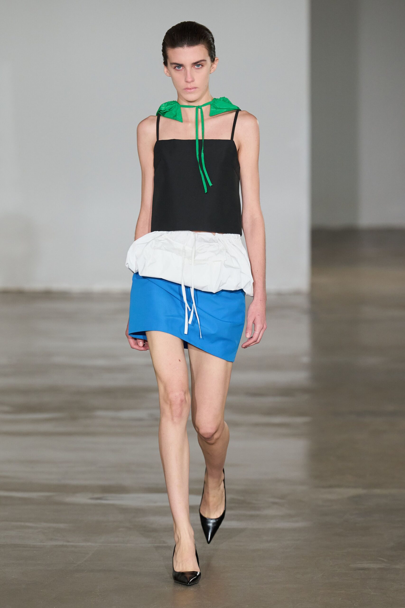 Bright green nylon hoodlet, tied close with two narrow cords. Black square-neck camisole with thin straps and a blunt cropped hem. White puffed peplum cinched by dangling drawstrings over a cobalt mini. Black patent pumps with a sharp pointed toe. Walking across a museum atrium with a rain shell tied at the throat.