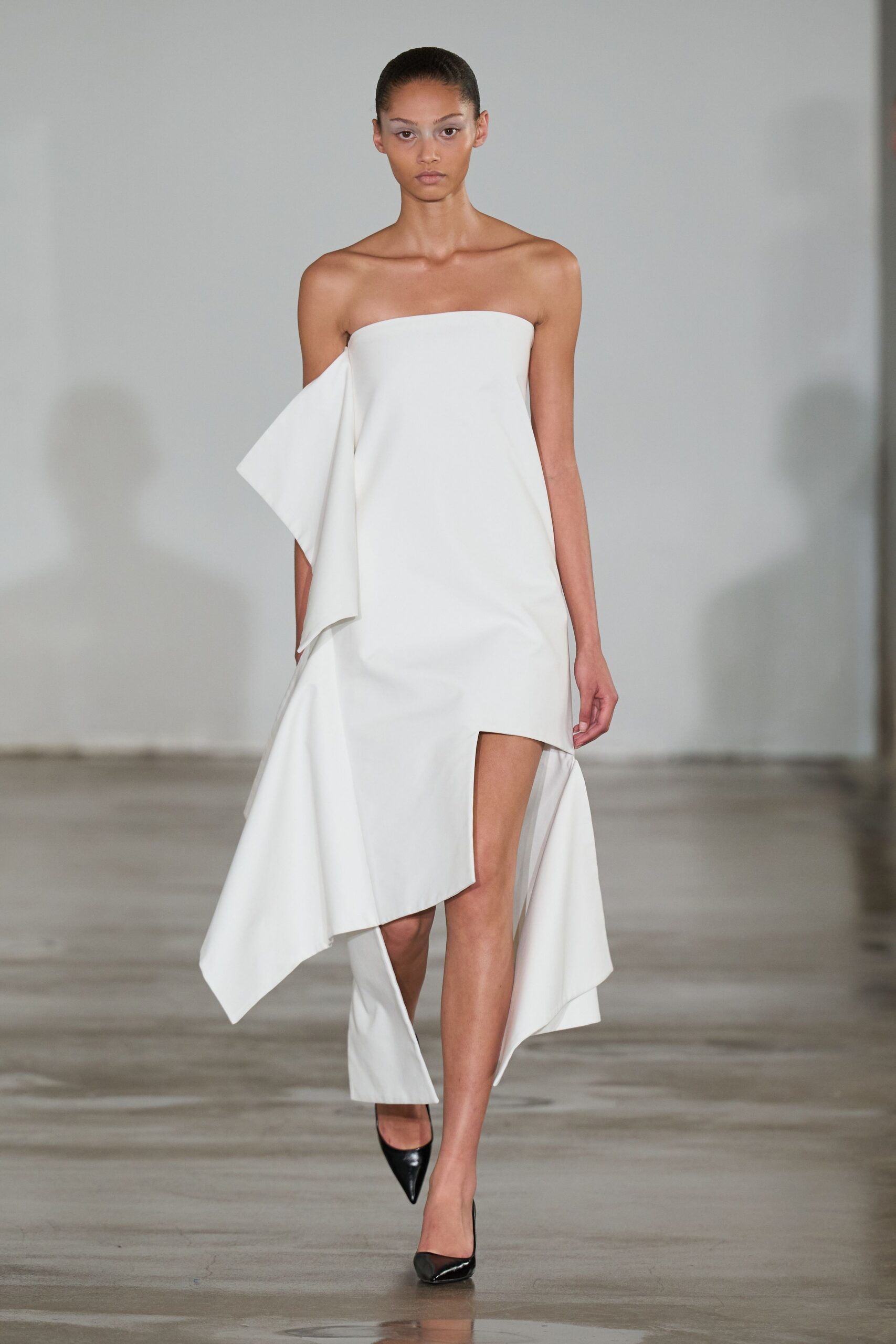 Strapless chalk-white dress with a folded side panel and jagged handkerchief hem. Black patent pumps with needle-sharp pointed toes. For crossing a museum atrium while the florist is still unpacking crates.