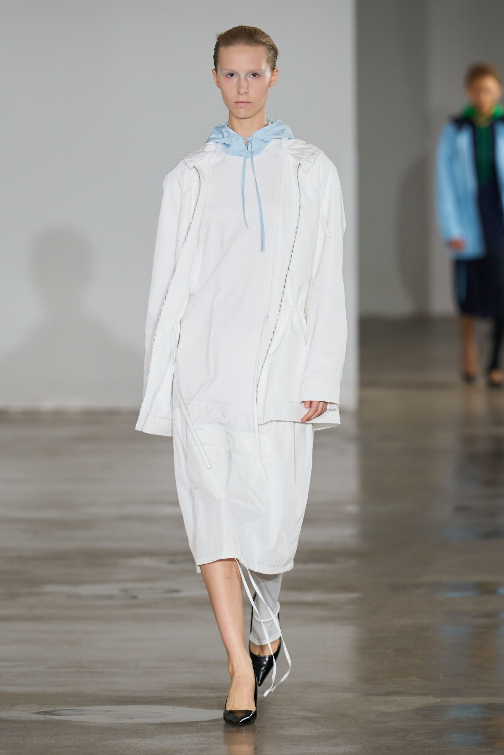 Powder-blue hood peeking out, tied with thin cord at the throat. White technical anorak dress with exposed zips and a dropped drawcord hem. Pale grey leggings under the uneven hem. Black patent pumps, sharply pointed. Walking the seawall before rain, map folded into a damp pocket.